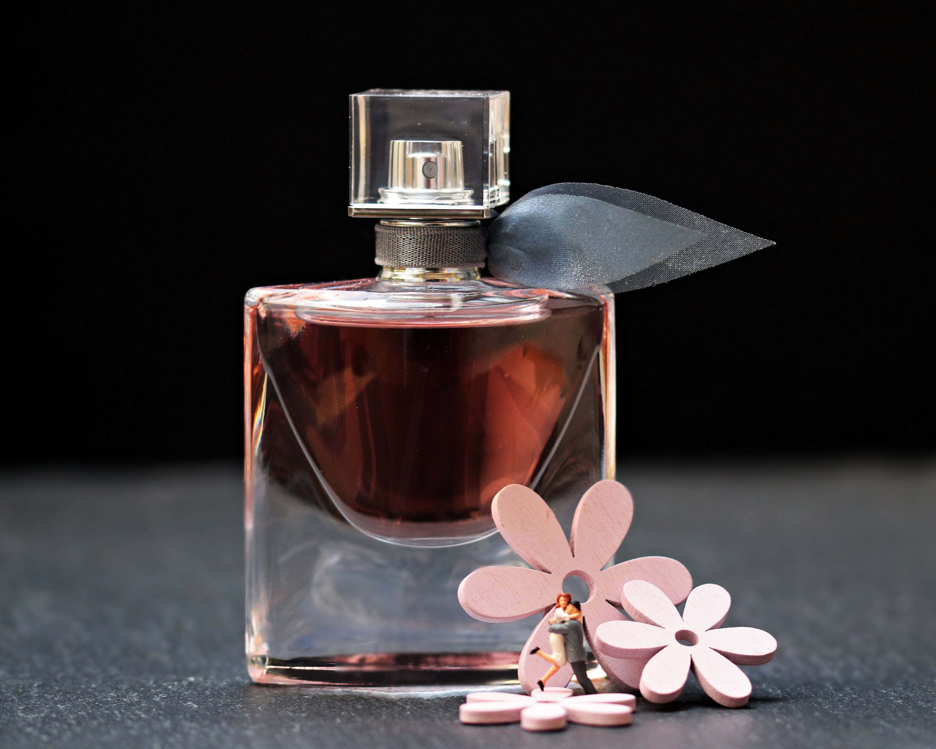 Floral Fragrance 桌布 壁紙 — 一個時尚的玻璃香水瓶，內有琥珀色液體和灰色絲帶，放在深色表面上，旁邊有粉紅色紙花和一個小情侶公仔。