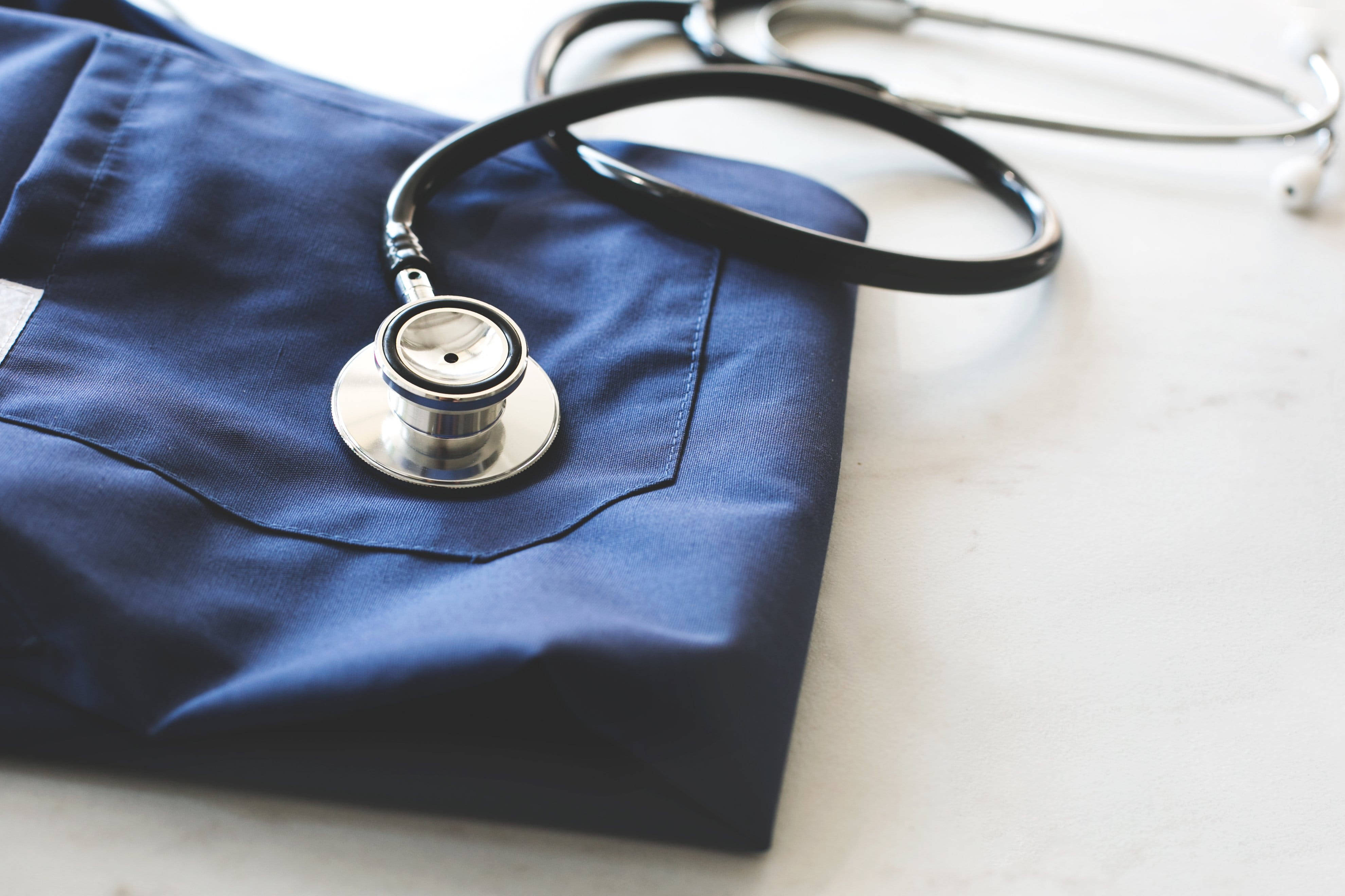 Stethoscope 桌布 壁紙 — 一個時尚的聽診器放在摺疊好的海軍藍色醫用刷手服上，置於潔白的表面上。