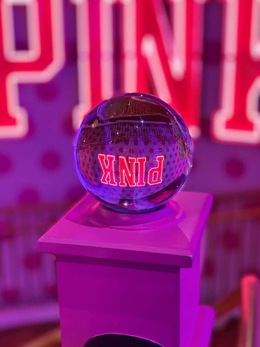 Victoria's Secret 桌布 壁紙 — 一個有光澤的紫色球體，上面有發光的紅色「PINK」文字，放置在相配的底座上，背景是模糊的霓虹燈招牌。