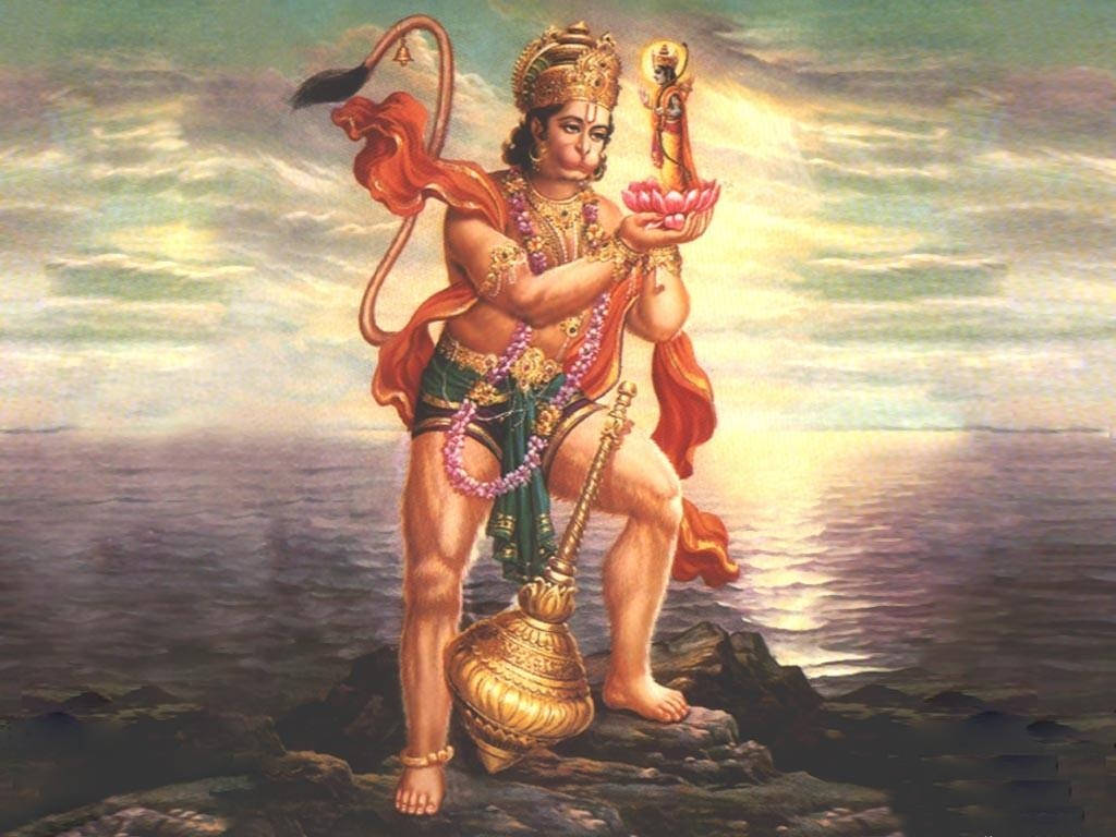 Hanuman 桌布 壁紙 — 一個有猴子尾巴的神聖身影站在岩石海岸上，手持蓮花和狼牙棒，背景是閃耀的夕陽大海。
