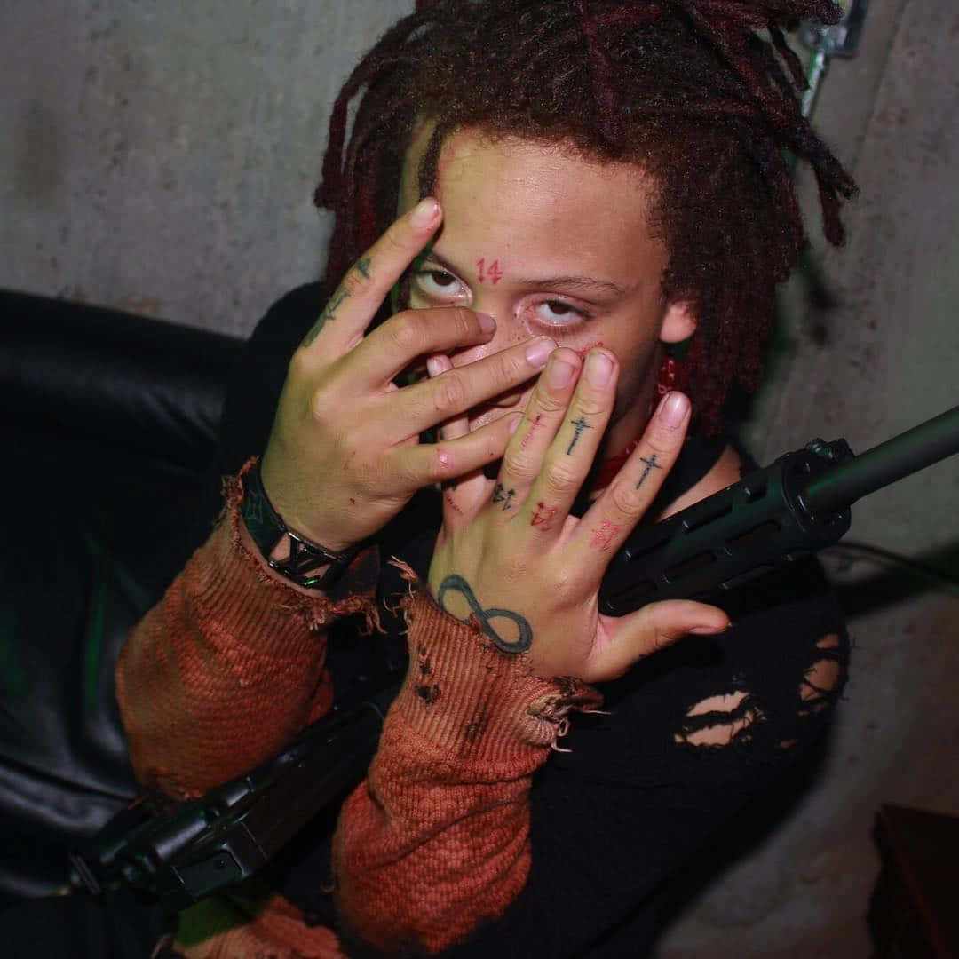 trippie redd 桌布 壁紙 — 一個有紅色辮子和紋身的人，在昏暗的房間裡用雙手遮住臉，同時拿著步槍。