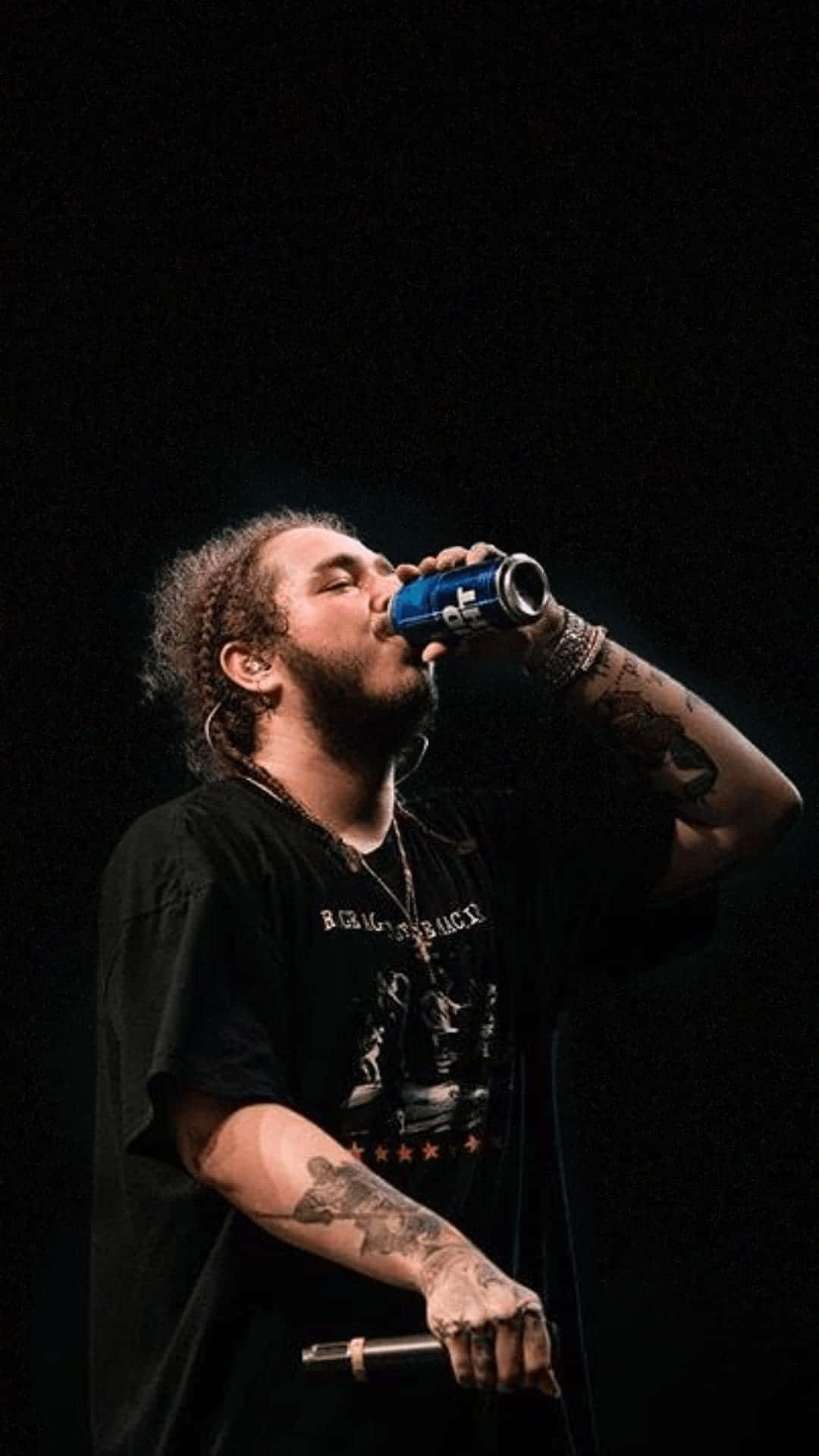 Post Malone 桌布 壁紙 — 一個有紋身、捲髮的男人在黑色背景的舞台燈光下喝藍色罐裝飲料。