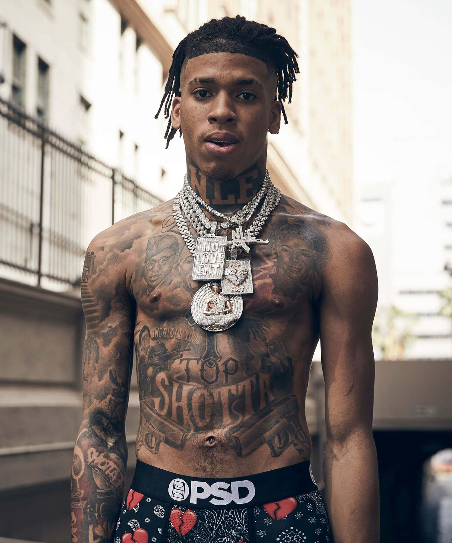 NLE Choppa 桌布 壁紙 — 一個有紋身、留著辮子、戴著沉重銀鏈的男人，在都市小巷中赤裸上身站立。