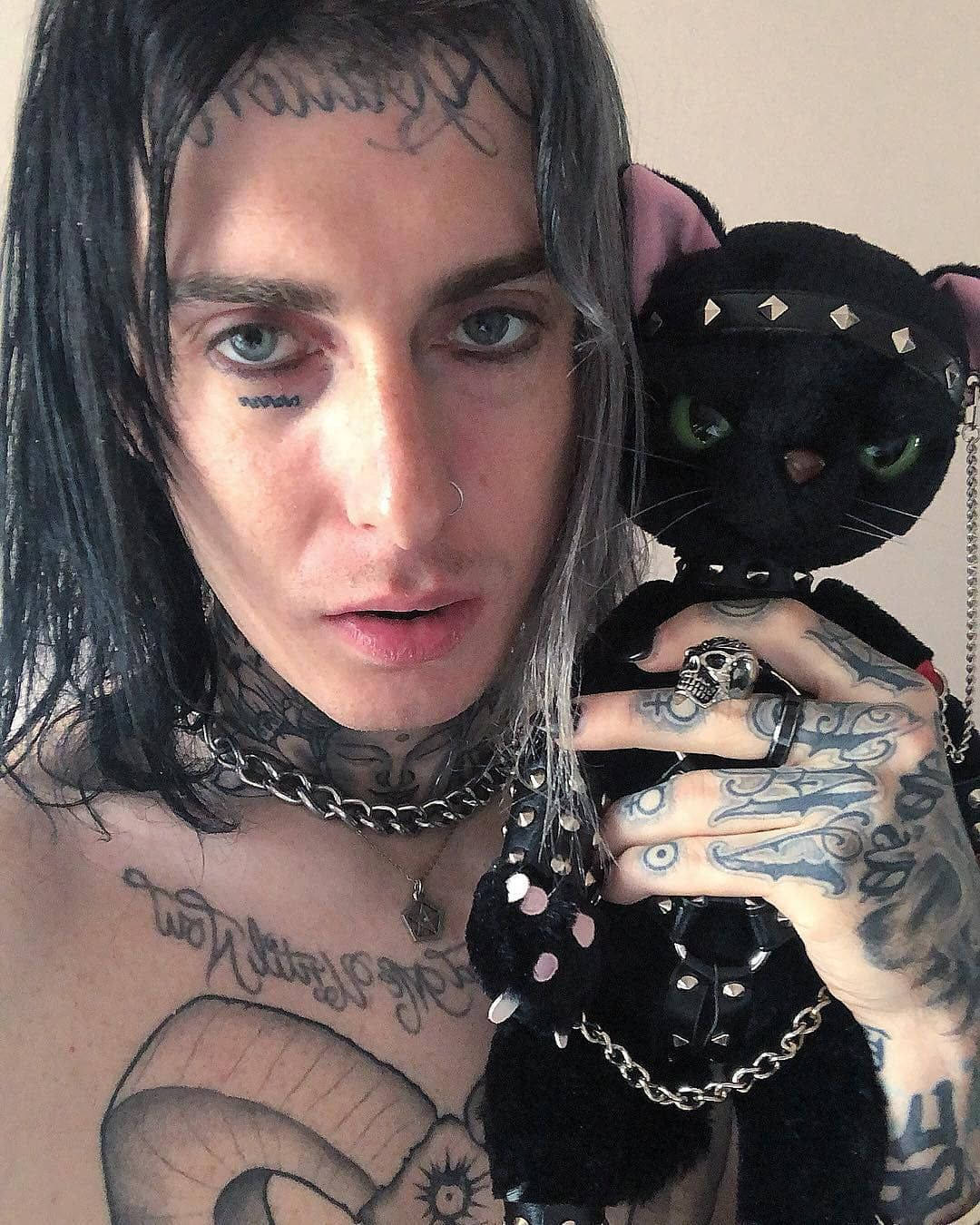 Ghostemane 桌布 壁紙 — 一個有紋身、皮膚蒼白、深色頭髮的人，拿著一隻黑色鉚釘貓咪毛絨玩具，靠著一面素牆。