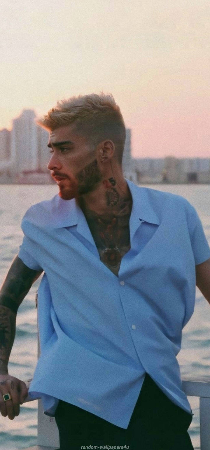 One Direction 桌布 壁紙 — 一個有紋身、身穿藍色恤衫嘅男人喺日落時企喺水邊，背後係城市天際線。