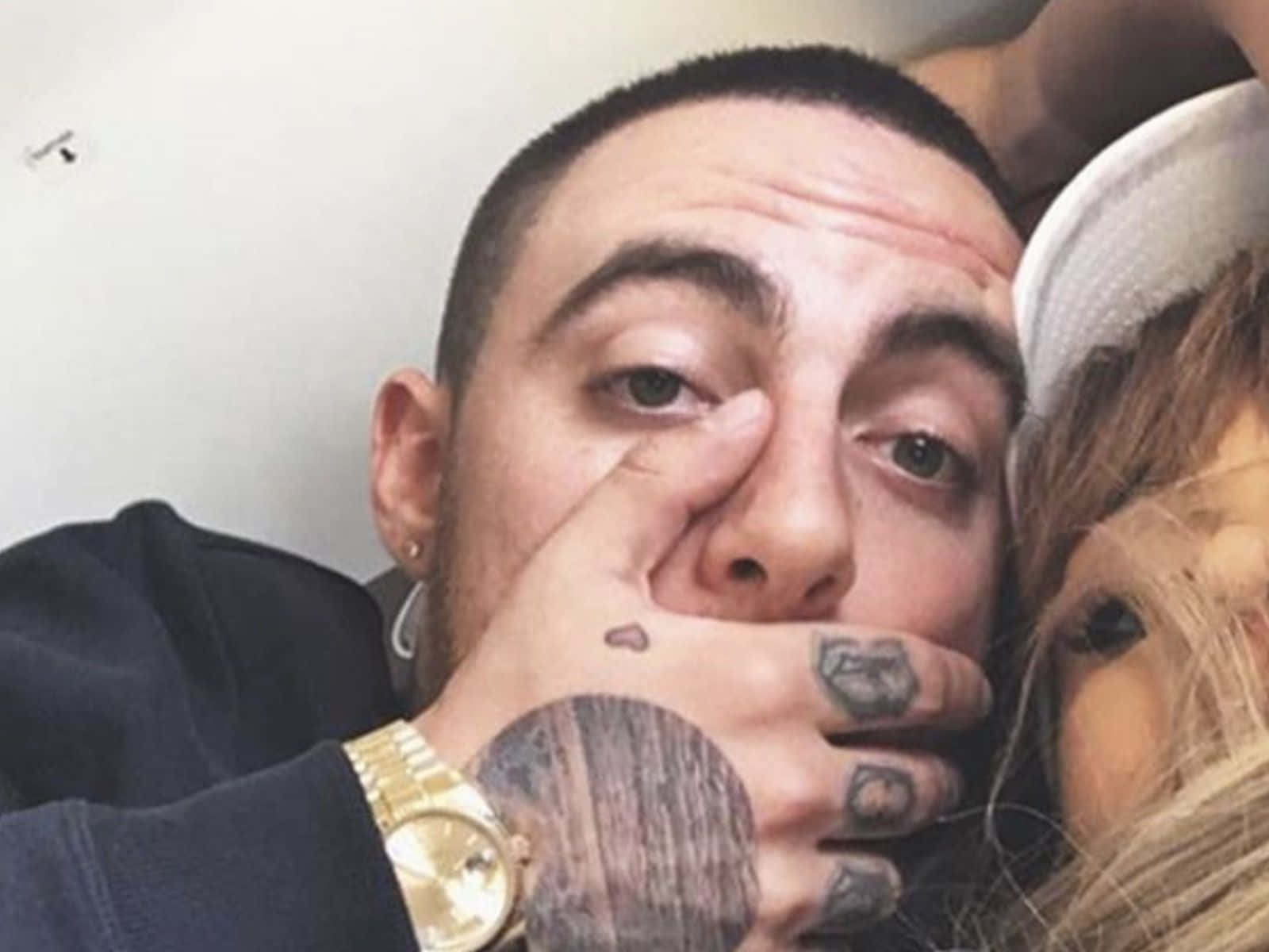 Mac Miller 桌布 壁紙 — 一個有紋身和戴金錶的男人，在擁抱一個戴白帽的女人時掩住自己的嘴。