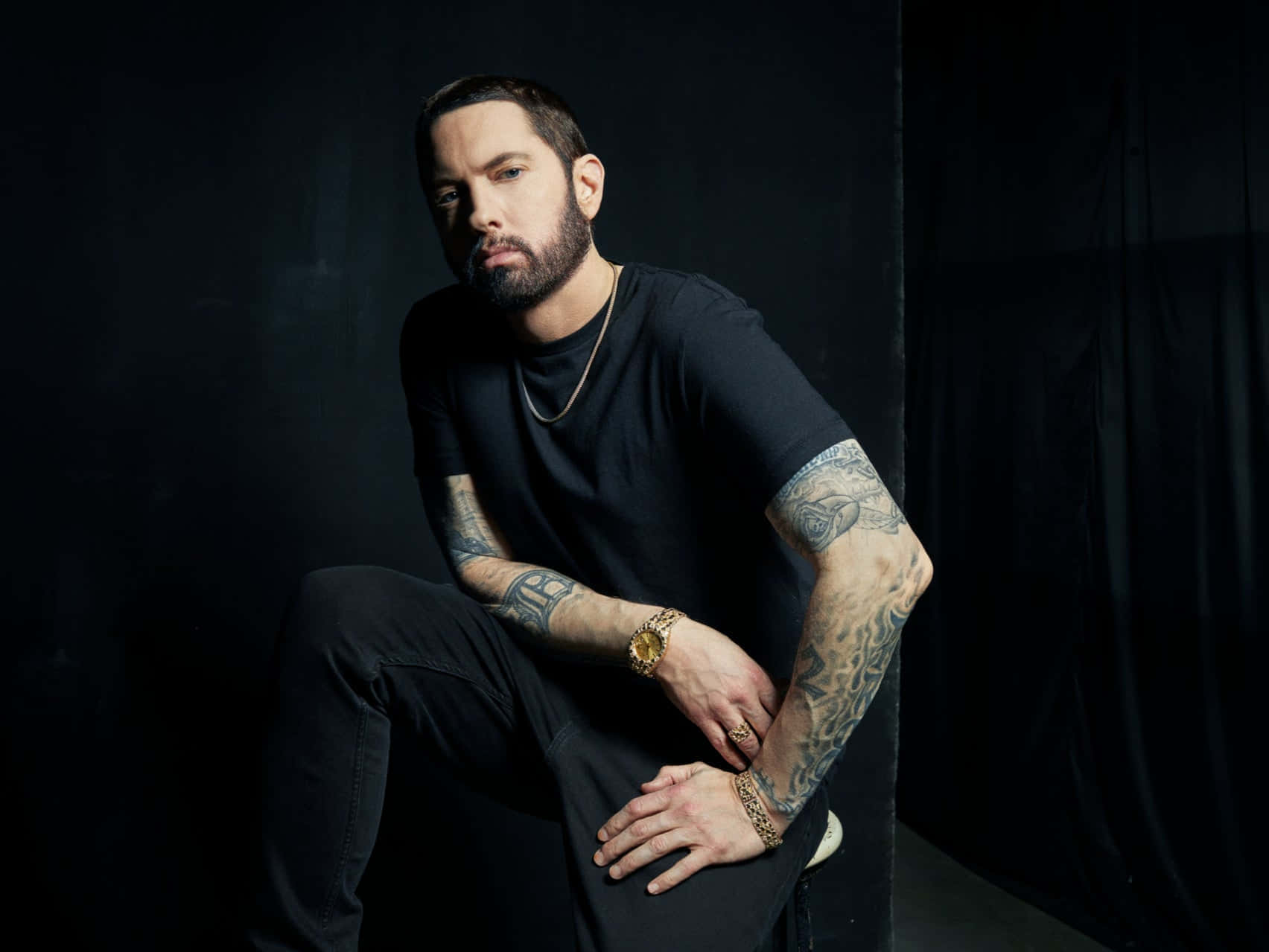 Eminem 桌布 壁紙 — 一個有紋身和戴金錶的男人坐在一個由戲劇性聚光燈照亮的黑暗工作室中。