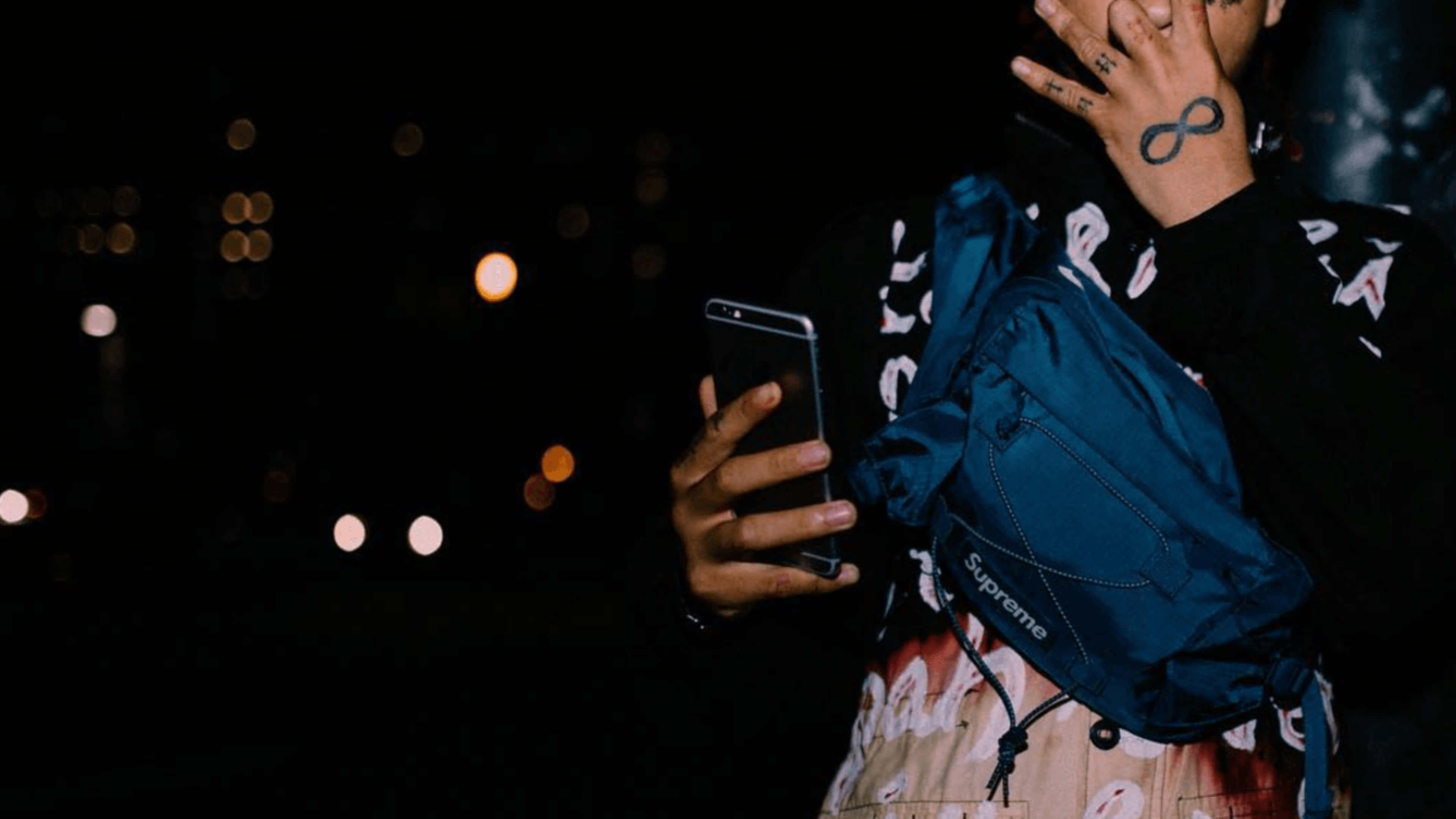 trippie redd 桌布 壁紙 — 一個有紋身嘅人喺模糊嘅夜景同發光嘅散景燈光背景下，揸住一部電話同一個藍色Supreme袋。