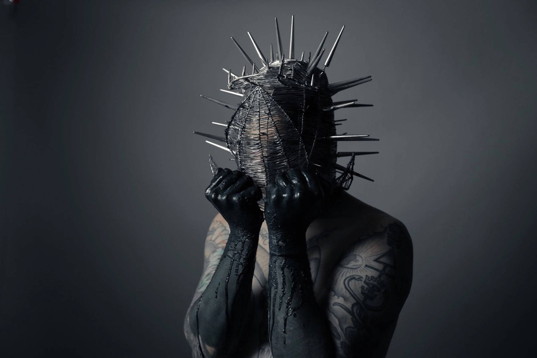 Ghostemane 桌布 壁紙 — 一個有紋身手臂的人戴著帶刺的金屬面具，並將其緊握在深灰色工作室背景前。