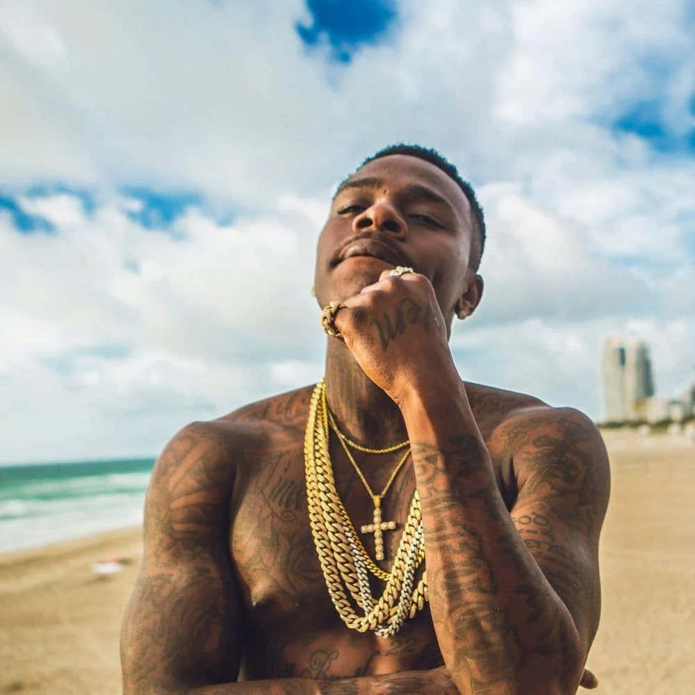 Dababy 桌布 壁紙 — 一個有紋身的男人戴著金鏈，在陽光明媚的海灘上仰望，背後是海洋和城市天際線。