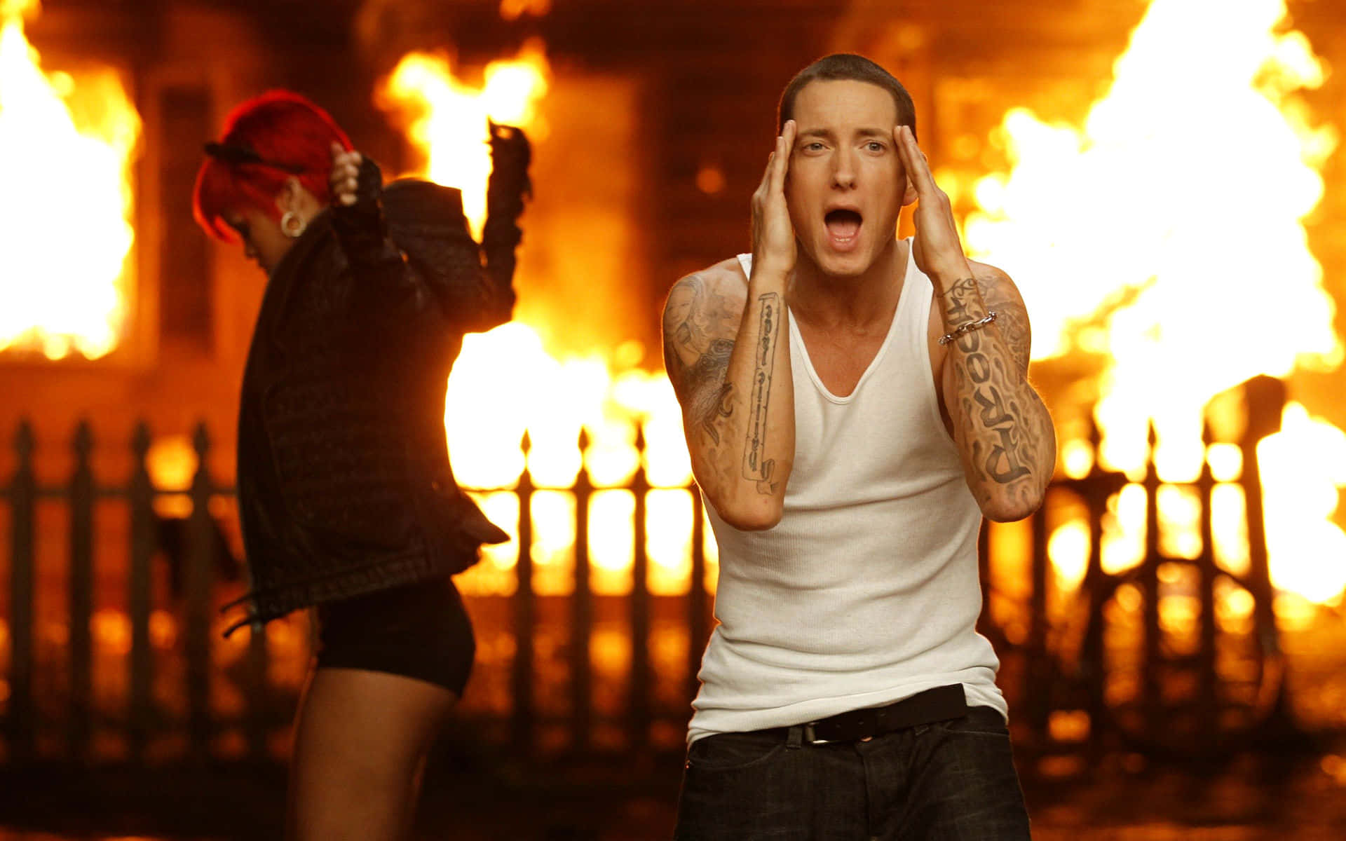 Eminem 桌布 壁紙 — 一個有紋身的男人驚恐地尖叫，火焰吞噬了他身後的圍欄，而一個紅髮女人在背景中跳舞。