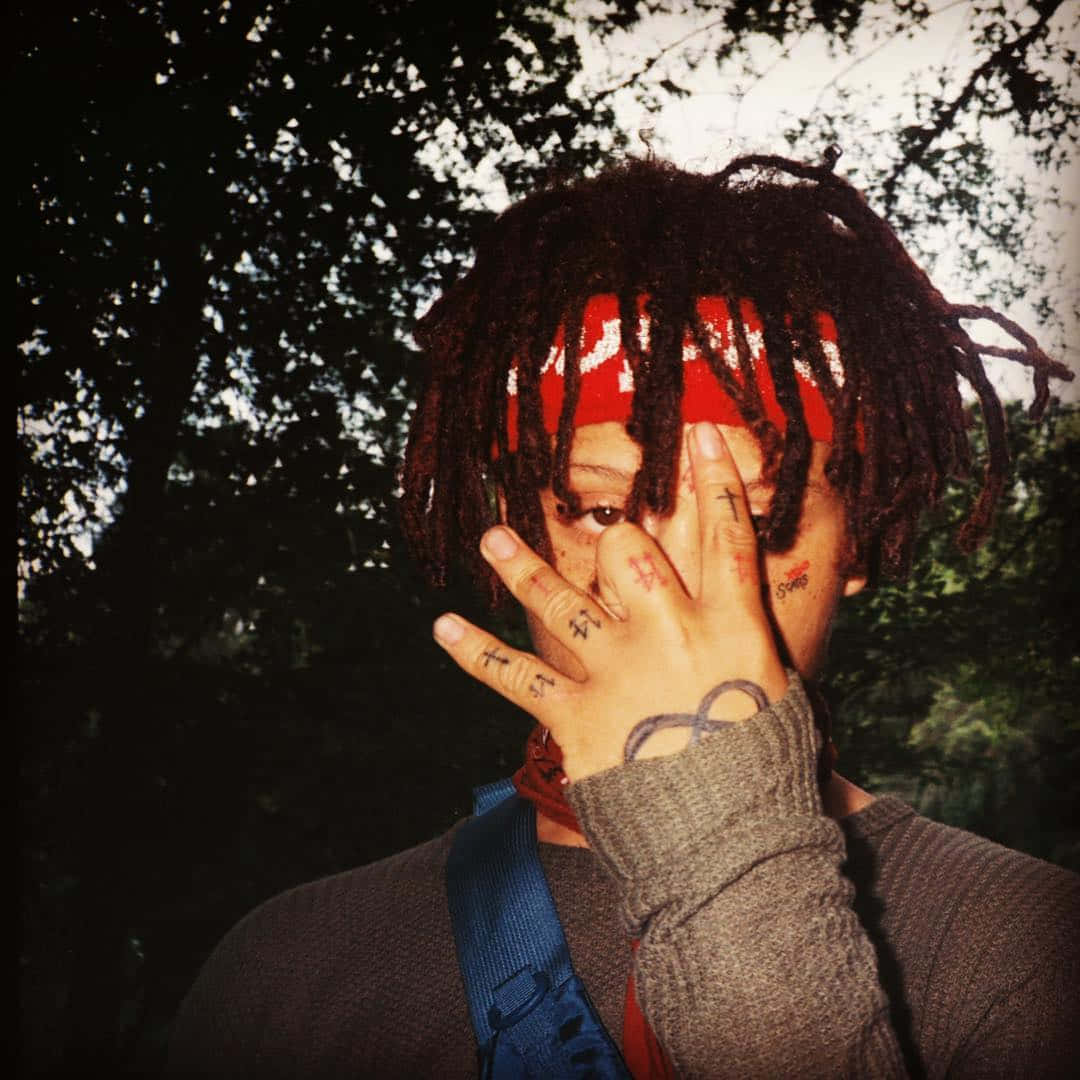 trippie redd 桌布 壁紙 — 一個有辮子頭和紋身的人站在樹林地區，用手遮住半邊臉。