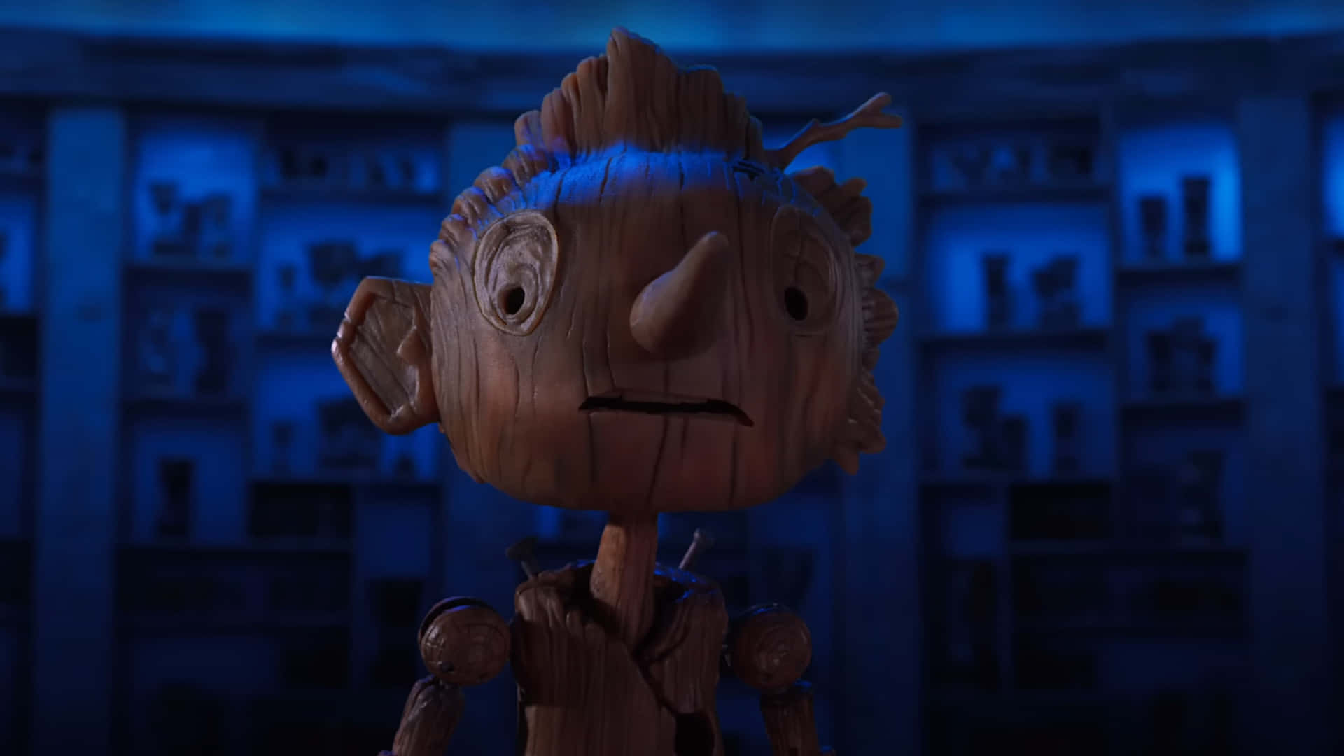 Guillermo Del Toro's Pinocchio 桌布 壁紙 — 一個木偶帶著憂慮的表情站在一個昏暗的藍色燈光房間裡，房間裡充滿了擺放著類似人物的架子。