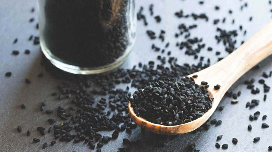 Black Cumin 桌布 壁紙 — 一個木勺盛滿光澤的黑芝麻，溢出到灰色表面上，旁邊有一個玻璃罐。