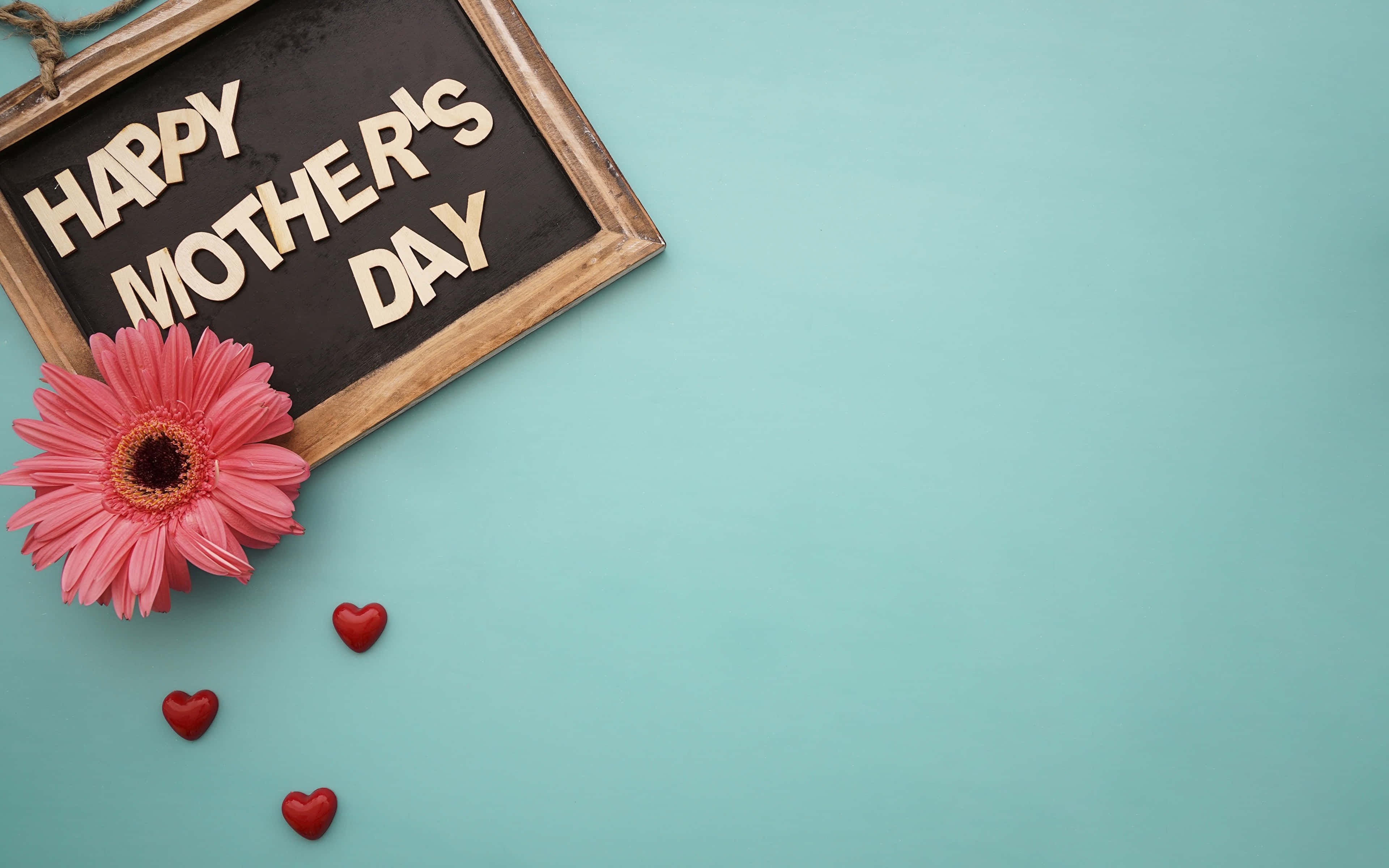 Happy Mothers Day 桌布 壁紙 — 一個木框「母親節快樂」標誌，在青綠色背景上有一朵粉紅色非洲菊雛菊和紅色心形。