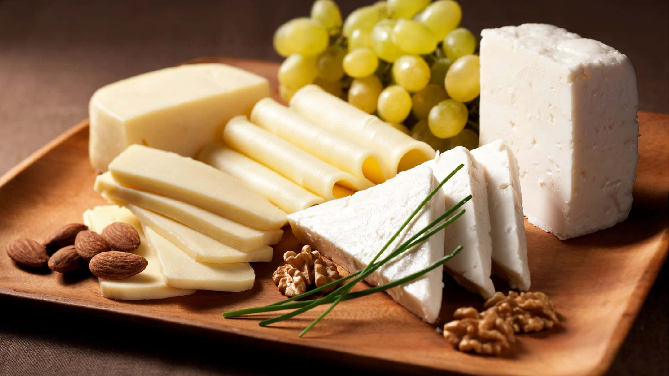 Cheese 桌布 壁紙 — 一個木盤在溫暖燈光下展示各式芝士、葡萄、堅果和香草。