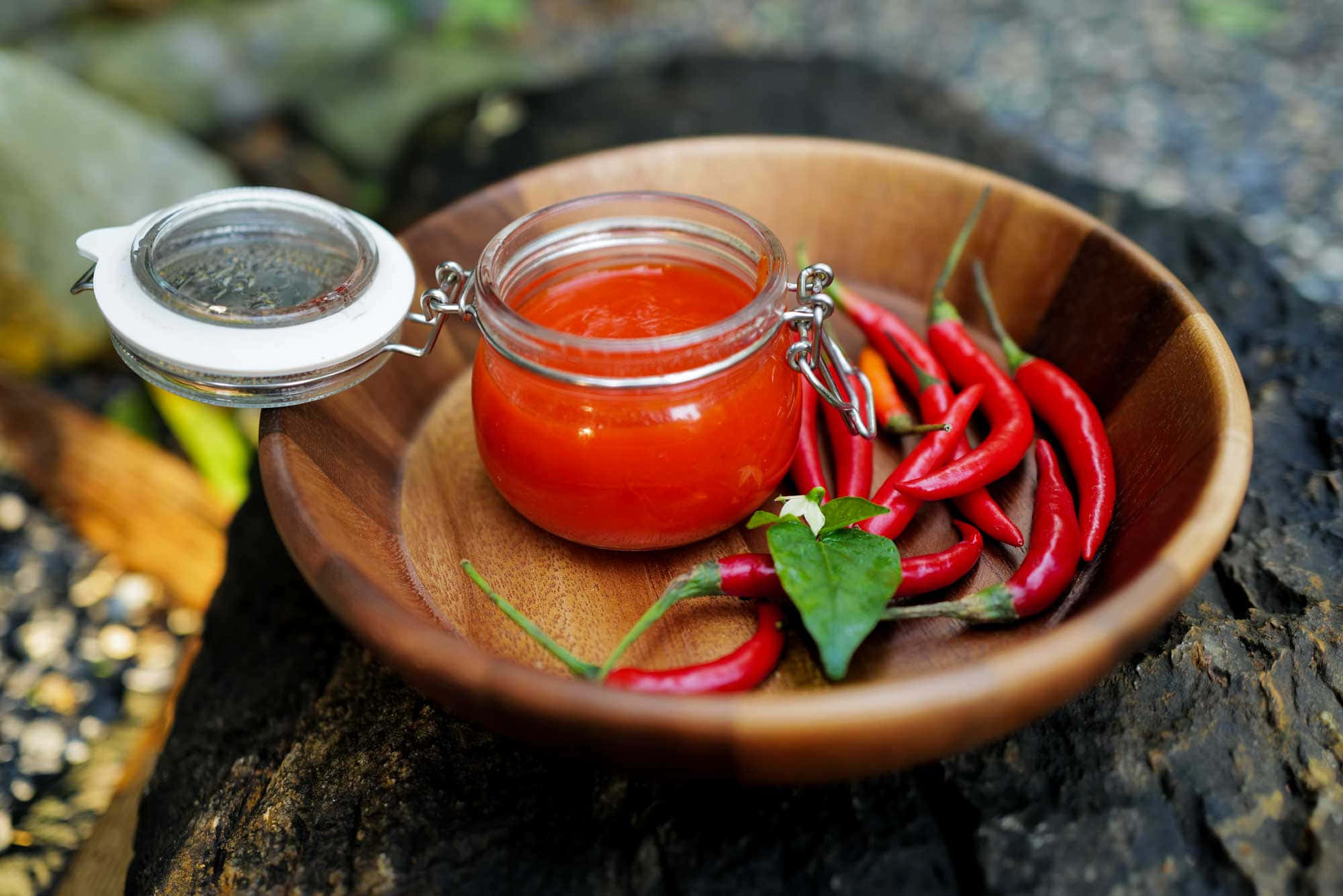Red Sauce 桌布 壁紙 — 一個木碗盛著一個玻璃罐的鮮紅辣椒醬，周圍是新鮮的紅辣椒和綠葉，放在質樸的石頭表面上。