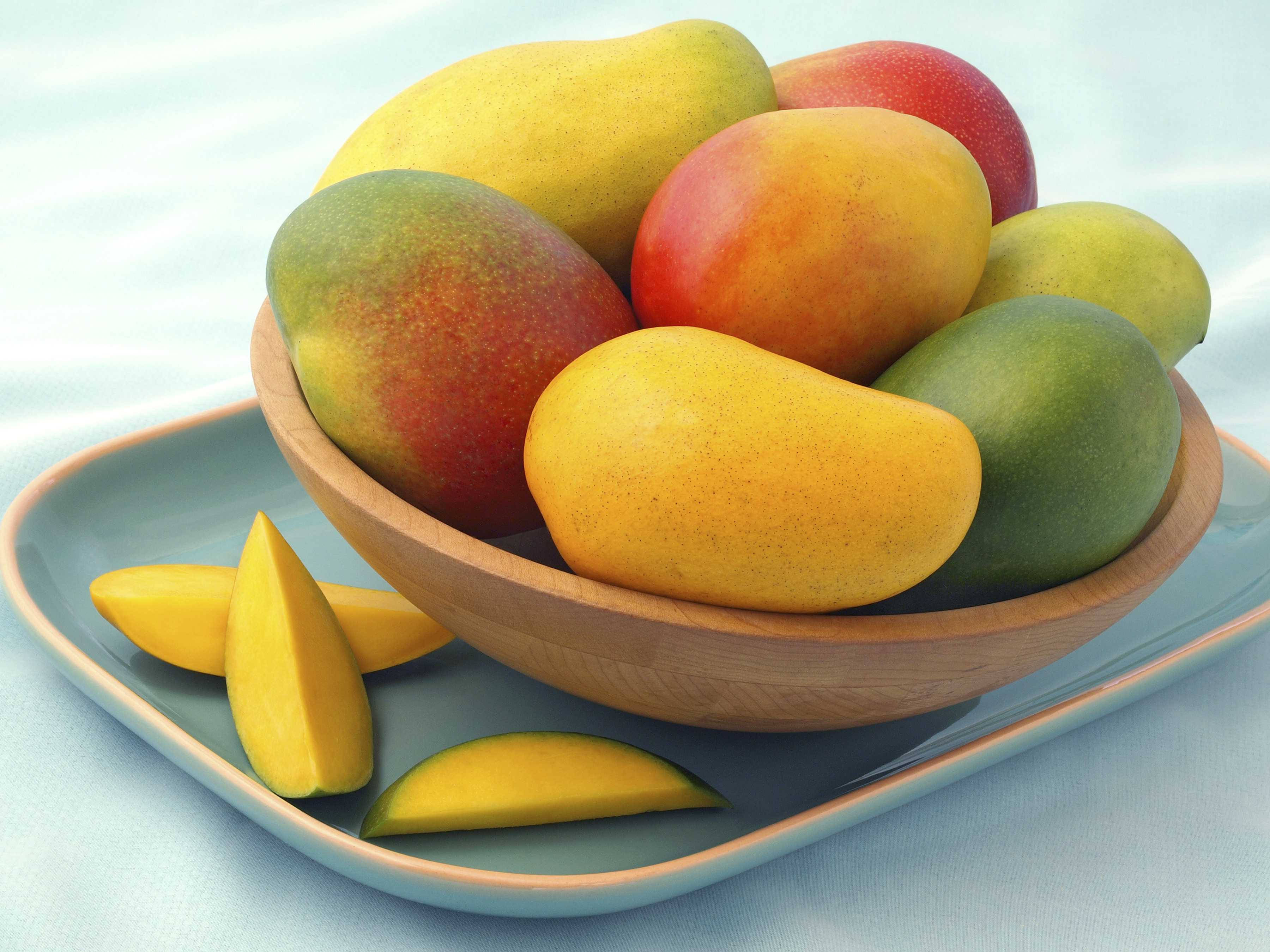 Mango 桌布 壁紙 — 一個木碗裝滿了黃色、紅色和綠色色調的成熟芒果，放在淺藍色托盤上，旁邊有切片芒果。