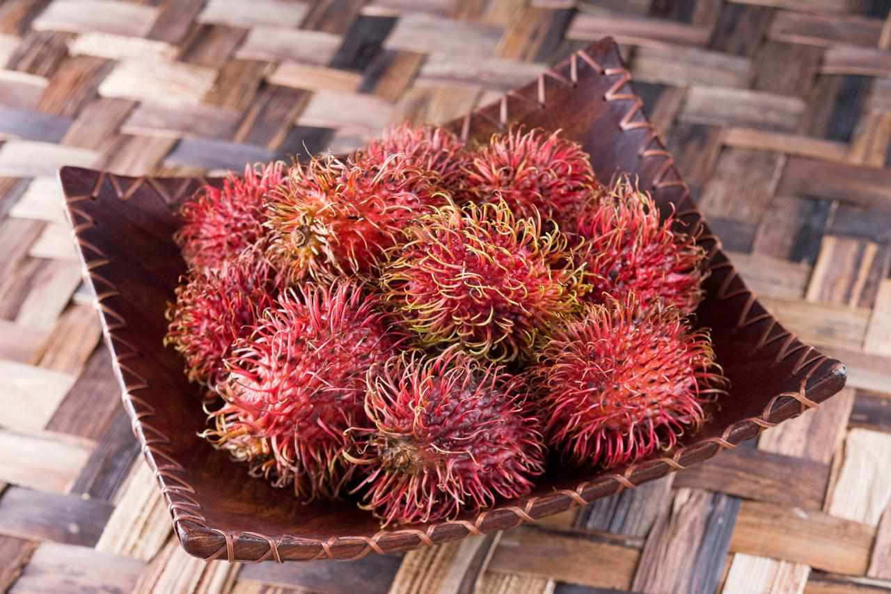 Red Fruit 桌布 壁紙 — 一個木碗裝滿鮮紅色的紅毛丹，放在一個編織竹蓆上，展現出複雜的紋理和溫暖的大地色調。