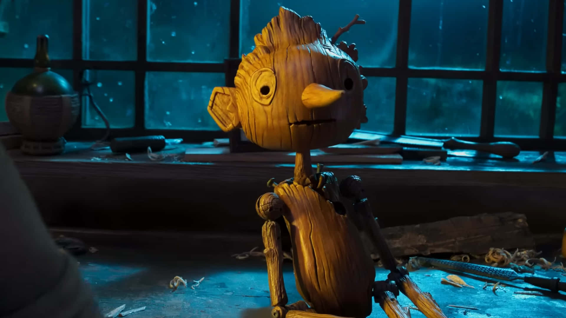 Guillermo Del Toro's Pinocchio 桌布 壁紙 — 一個木製動畫角色，有樹枝四肢，獨自坐在昏暗的工作室中，月光從窗戶照進來。