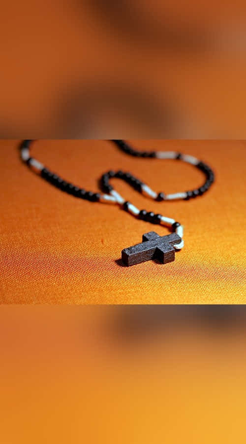 Sacrament 桌布 壁紙 — 一個木製十字架吊墜掛在黑白色珠鏈上，放在溫暖的橙色紋理表面上。