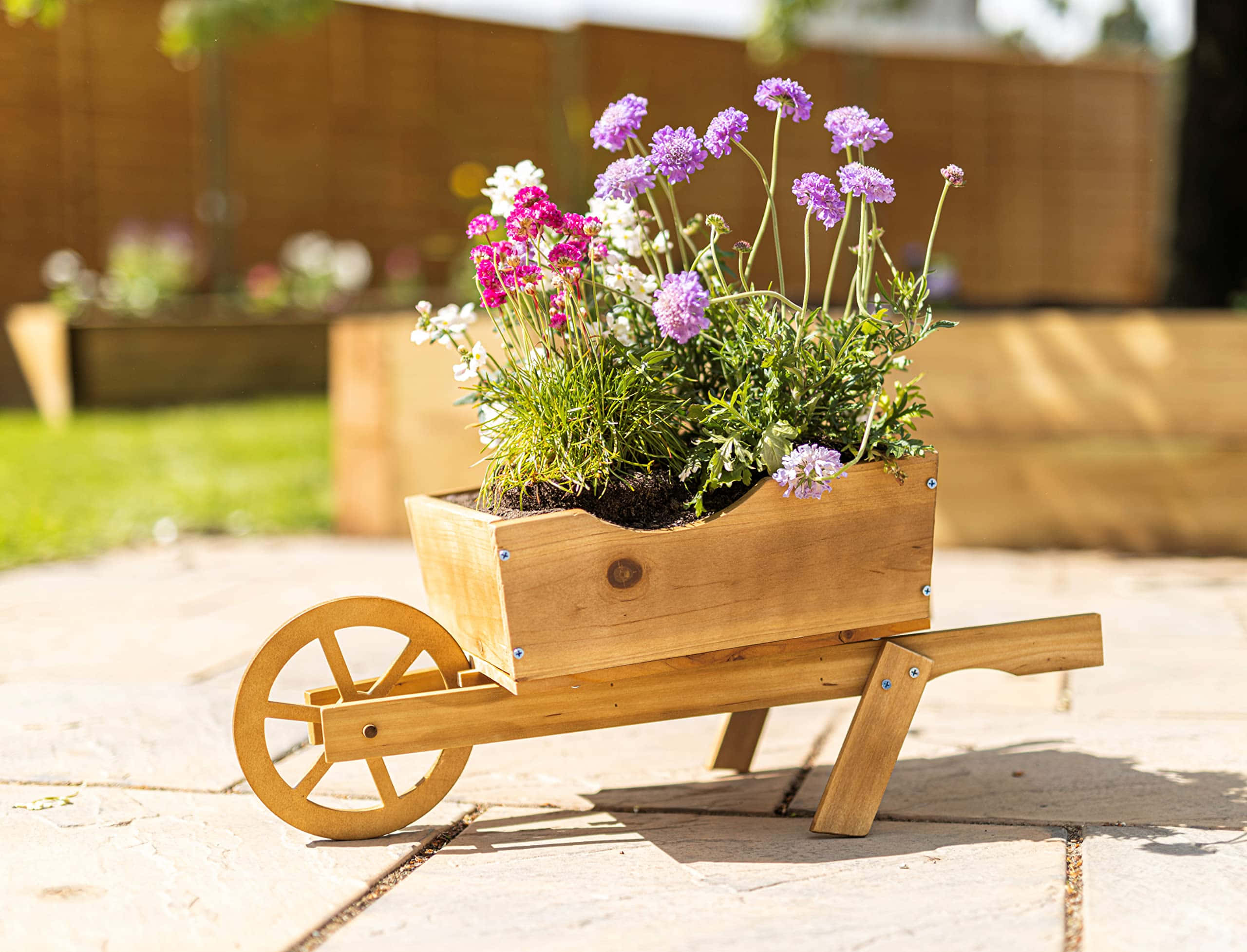 Flower Cart 桌布 壁紙 — 一個木製手推車花盆，滿載著紫色、粉紅色和白色的花朵，放置在陽光普照的石頭庭院中。