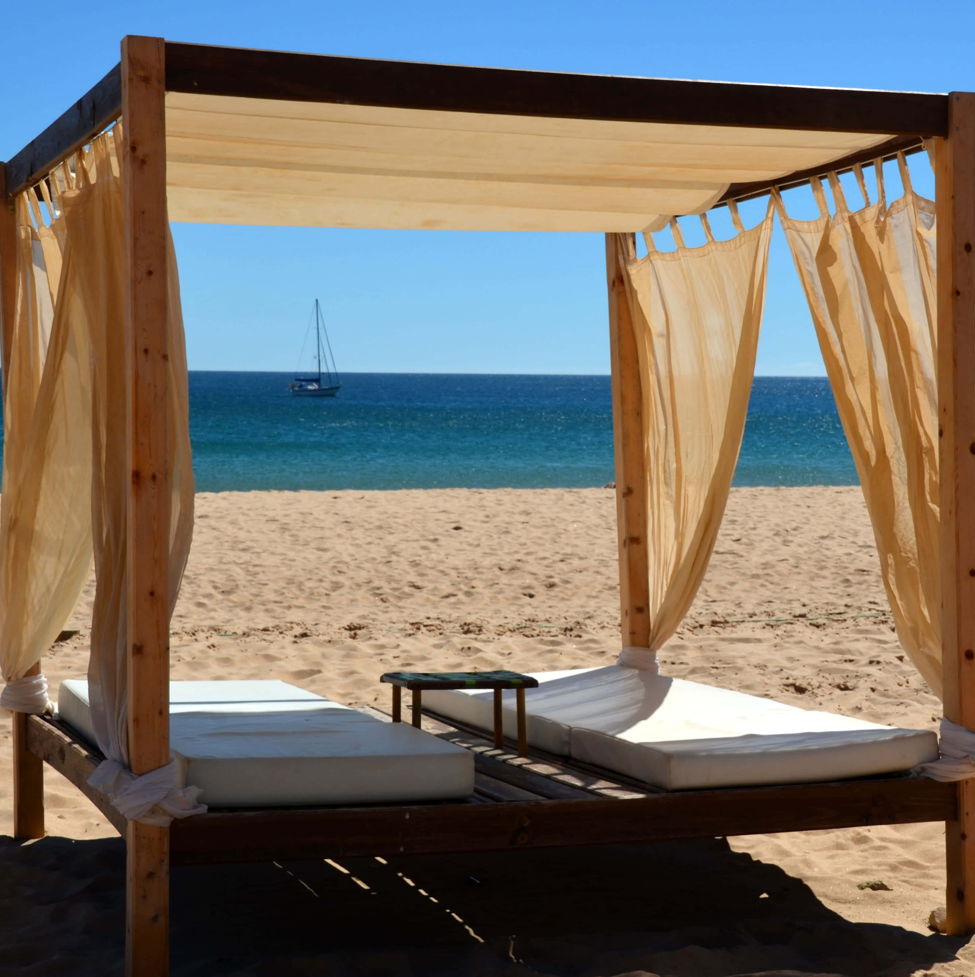 Beach Daybed 桌布 壁紙 — 一個木製涼亭配米色窗簾，框住沙灘上的兩張日光浴床，俯瞰碧綠海洋，一艘帆船在清澈藍天下。