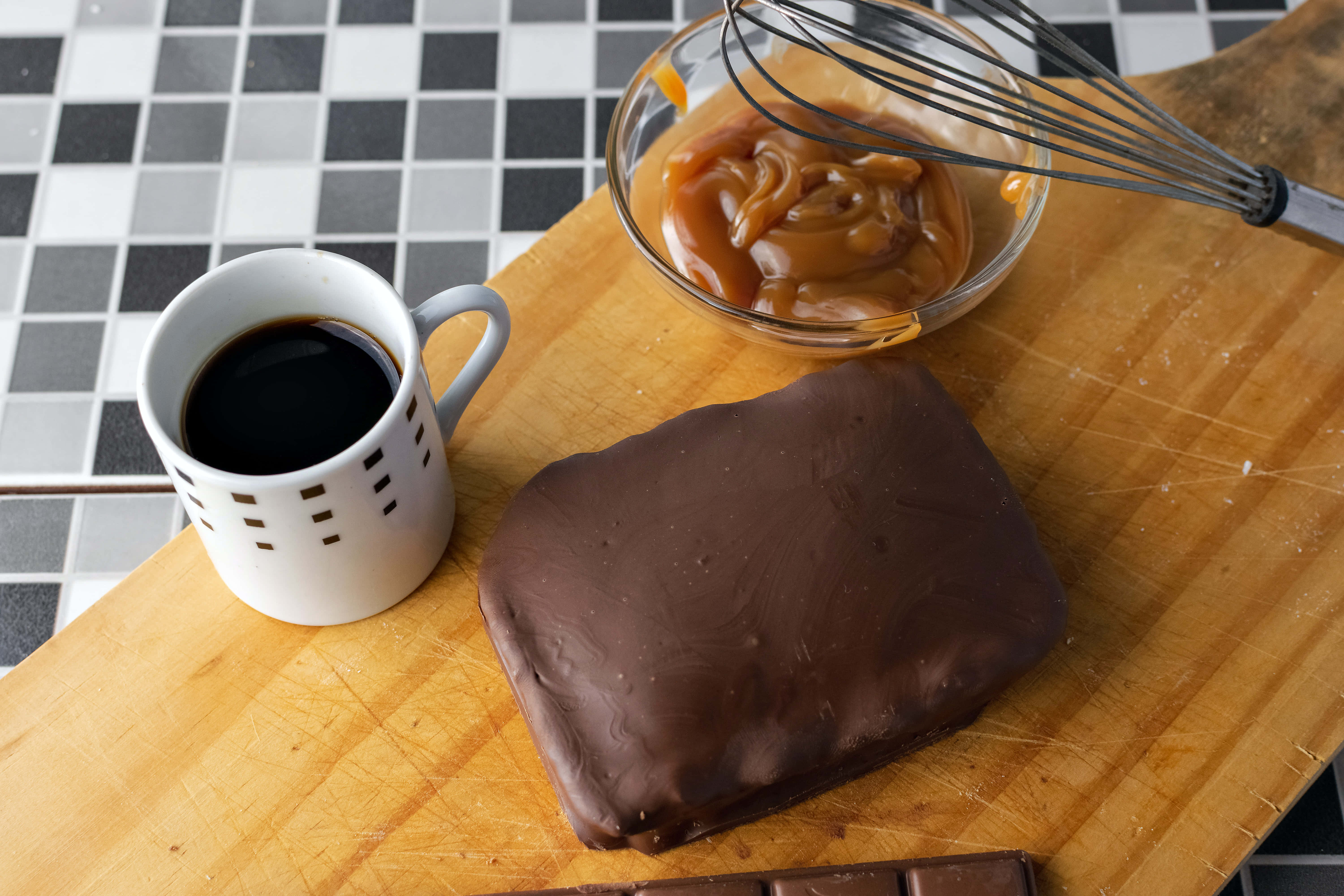 Caramel 桌布 壁紙 — 一個木製砧板上放著一塊朱古力焦糖糖果、一碗焦糖醬和一杯黑咖啡，置於格子瓷磚檯面上。