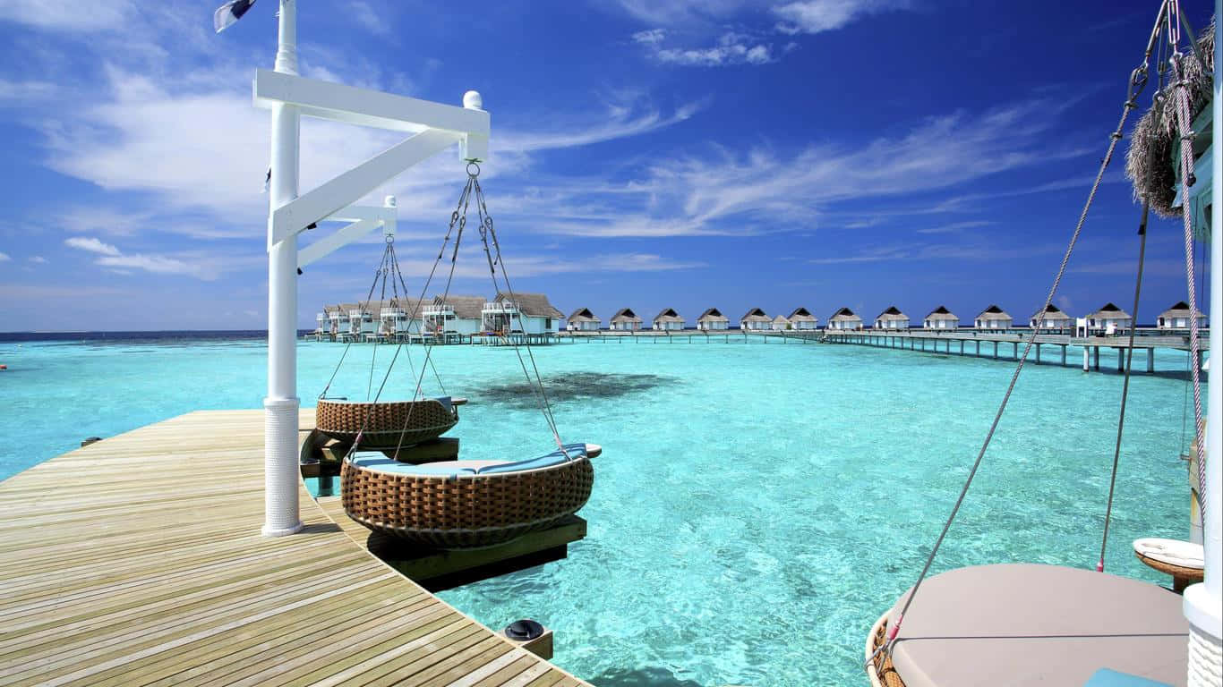 Maldives Island 桌布 壁紙 — 一個木製碼頭，掛著柳條吊椅，俯瞰著點綴著水上平房的碧綠海水，頭頂是明亮的藍天。