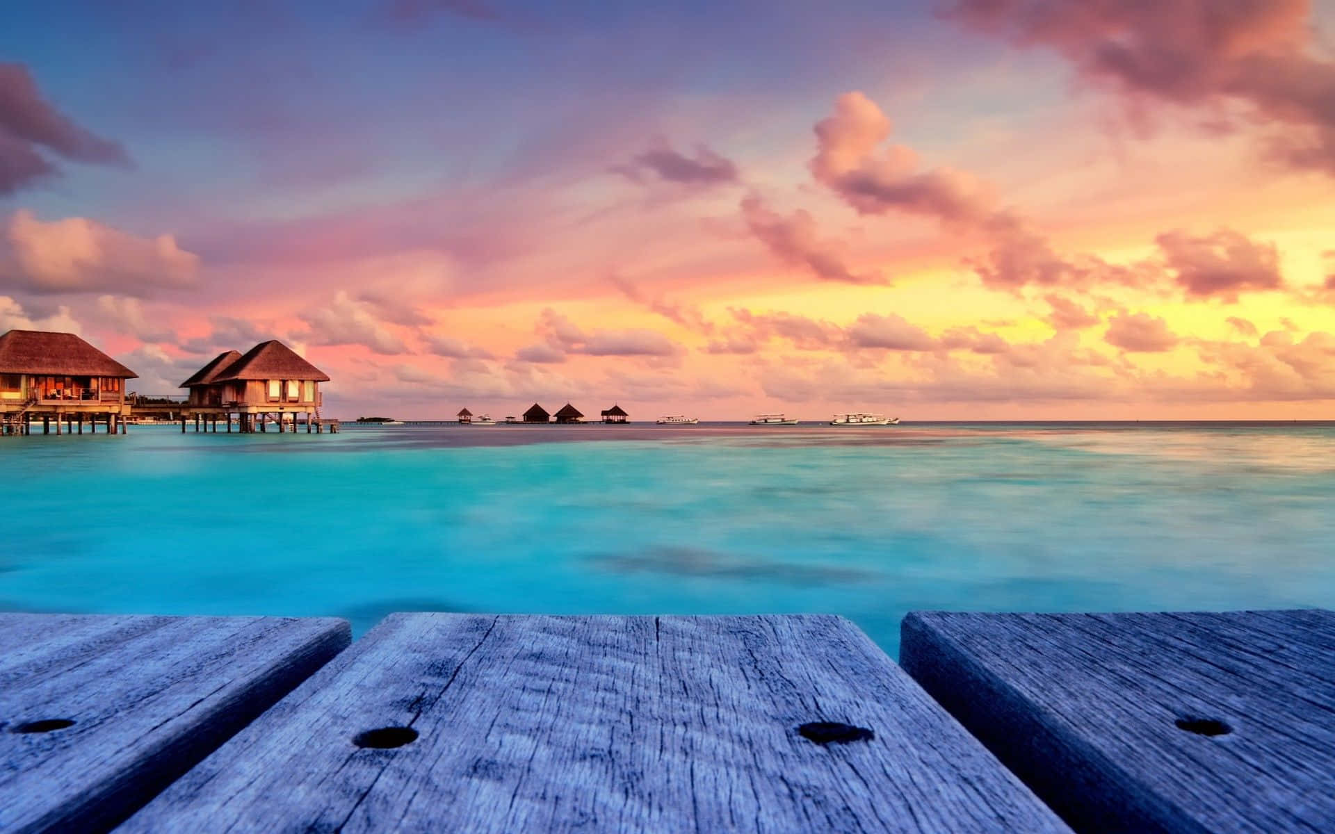 Maldives Beach 桌布 壁紙 — 一個木製碼頭框住了一個寧靜的熱帶日落，有粉彩雲和點綴著水上平房的碧綠海水。