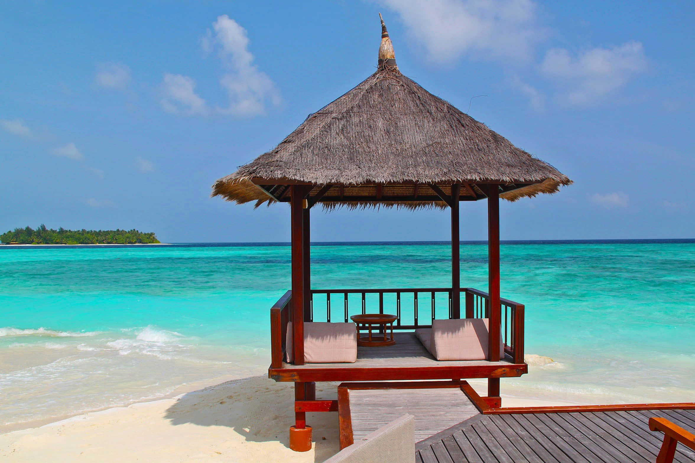 Beach Getaway 桌布 壁紙 — 一個木製茅草涼亭坐落在純淨的白色沙灘上，俯瞰著蔚藍的海水，上方是明亮的藍天。