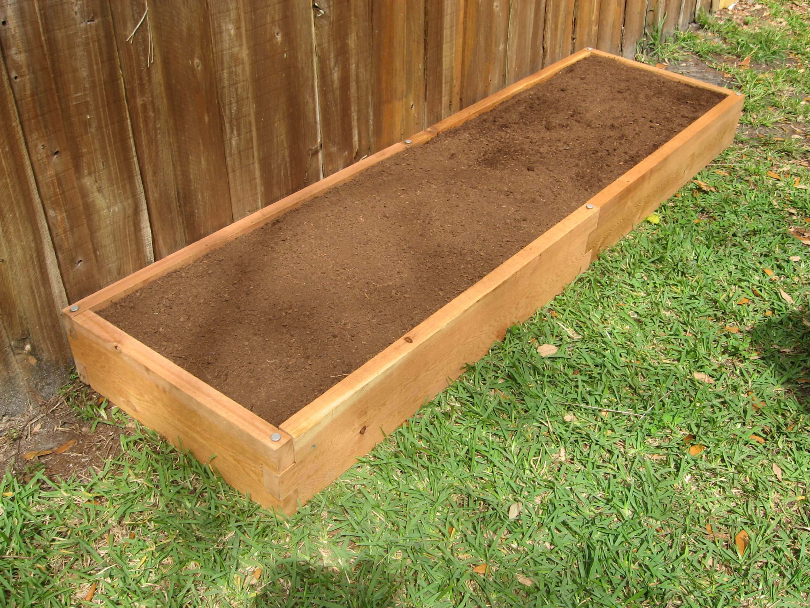 Garden Bed 桌布 壁紙 — 一個木製高架花圃裝滿了肥沃的棕色土壤，放在綠色草地上，旁邊是風化的木籬笆。