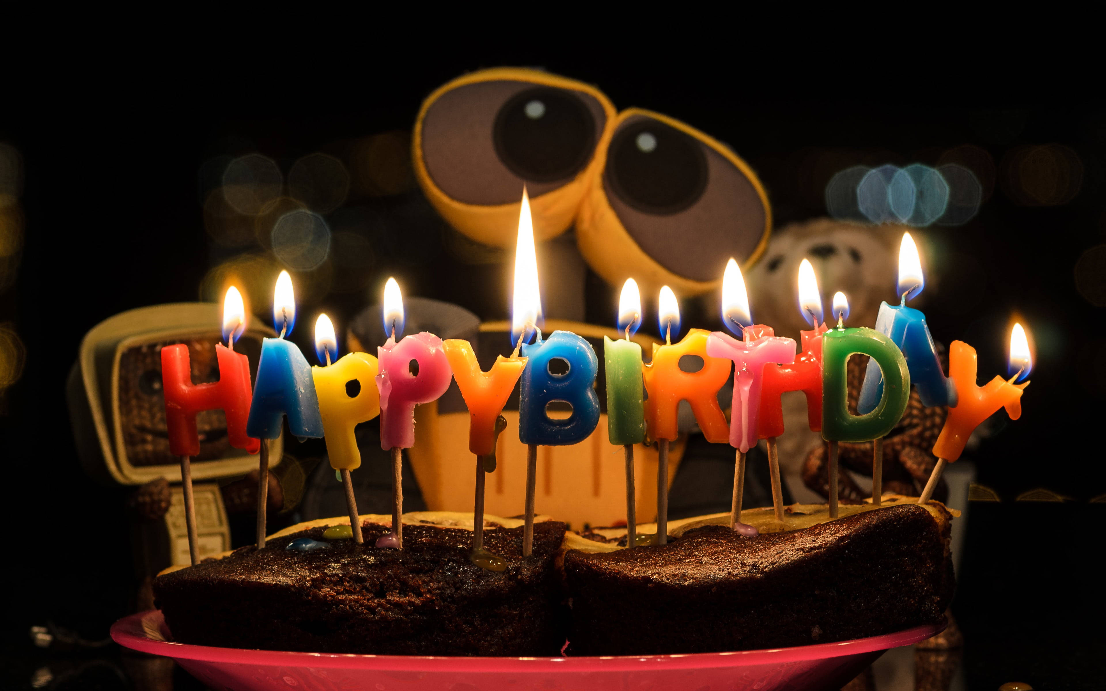 Birthday 桌布 壁紙 — 一個朱古力生日蛋糕，上面有色彩繽紛的「生日快樂」蠟燭，在黑暗的散景背景前，一個大眼睛機械人玩具前點燃。