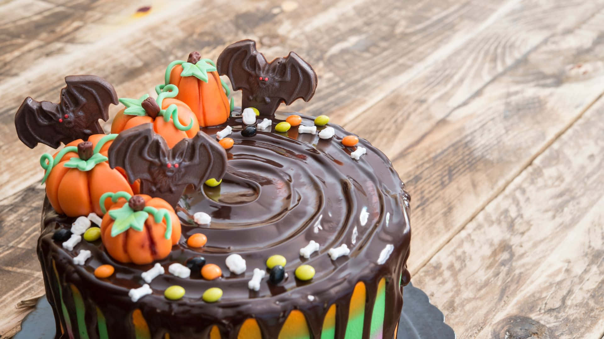 Halloween Cake 桌布 壁紙 — 一個朱古力萬聖節蛋糕，飾有橙色南瓜、朱古力蝙蝠和糖果骨頭，放在質樸的木桌上。