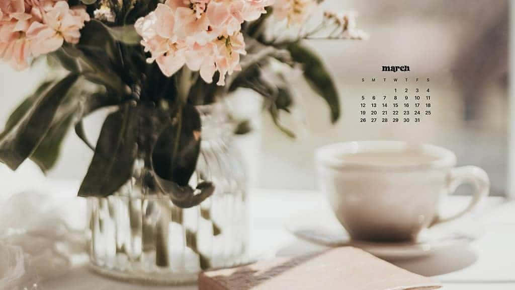 March Desktop 桌布 壁紙 — 一個柔和粉彩的三月日曆，旁邊放著一杯冒熱氣的飲品和一個插滿粉紅色花朵的花瓶，放在陽光灑落的桌子上。