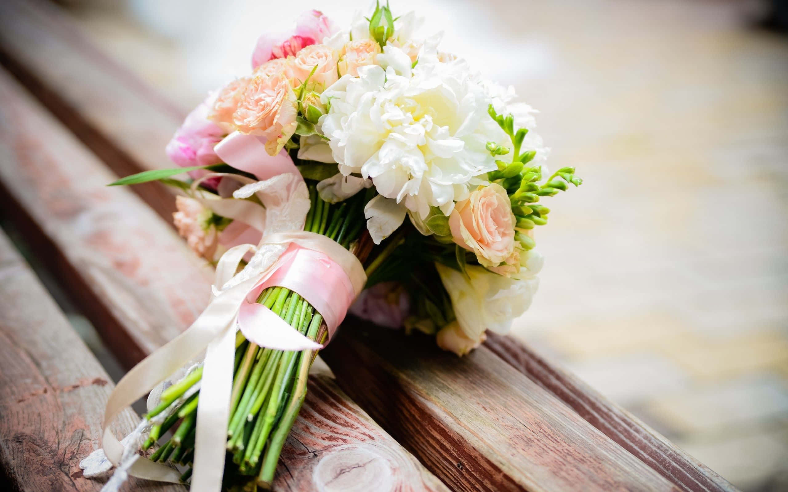 Wedding Flowers 桌布 壁紙 — 一個柔和粉彩的婚禮花束，由玫瑰和牡丹組成，繫上粉紅色絲帶，放在一張風化的木長椅上。