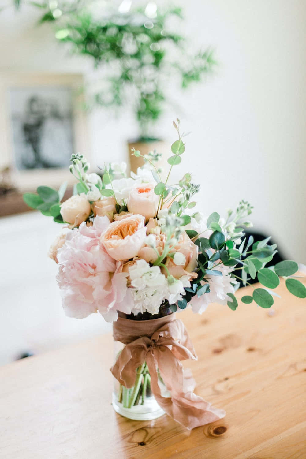 Floral Arrangement 桌布 壁紙 — 一個柔和粉彩花卉佈置，玻璃花瓶裡插著牡丹和尤加利葉，繫上灰玫瑰色絲帶，放在溫暖的木桌上。