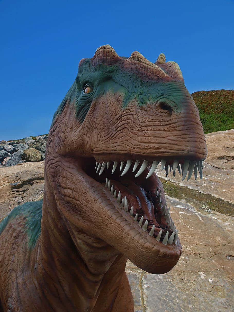 3d Dinosaur 桌布 壁紙 — 一個栩栩如生的暴龍雕像，帶有紋理的棕色和綠色皮膚，在岩石地形上對著晴朗的藍天咆哮。