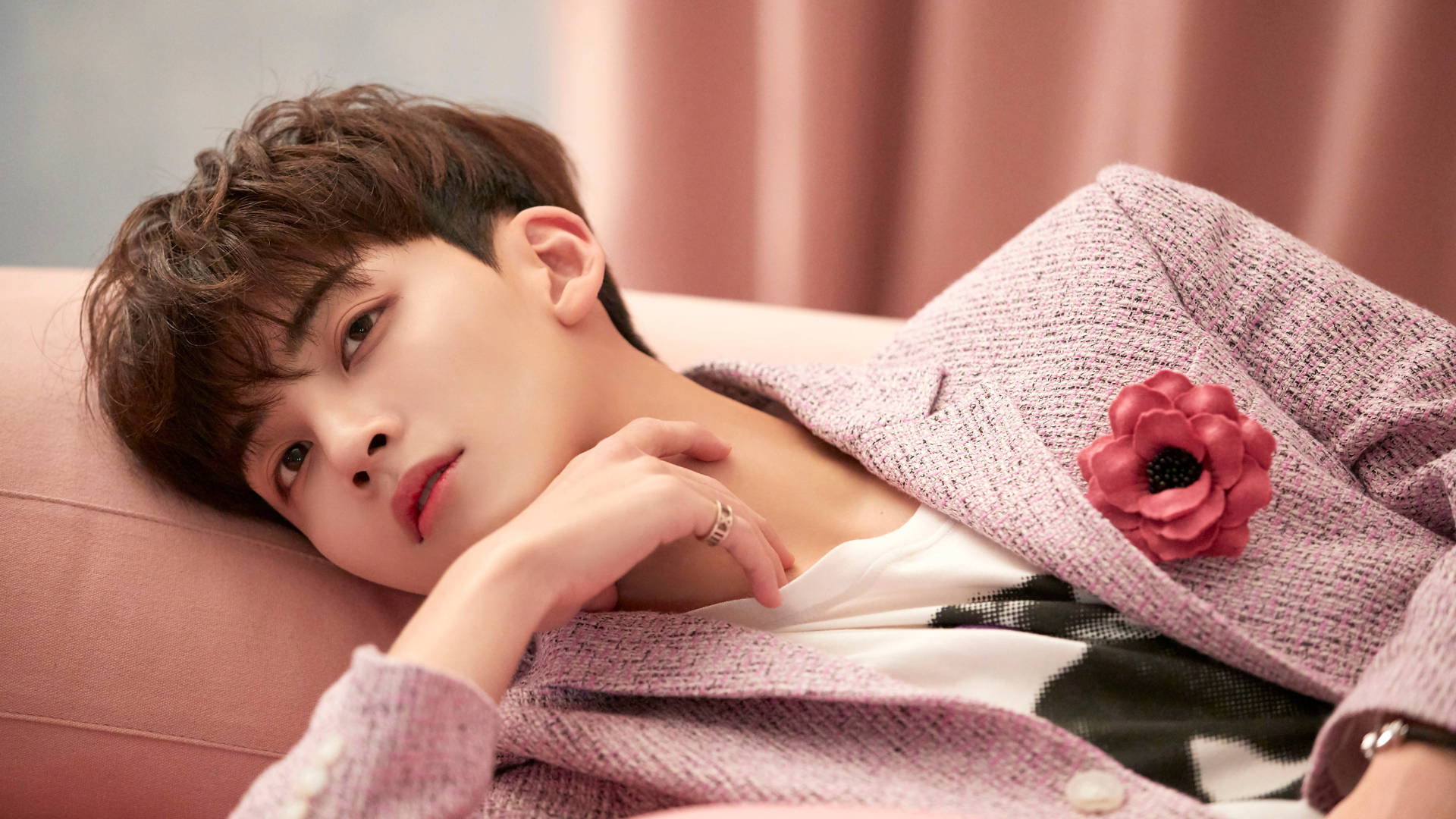 Yoon Jeonghan Fallin' Flower 壁紙 — 一個棕色亂髮的年輕男子將頭靠在粉紅色沙發上，身穿帶有紋理的粉紅色西裝外套，飾有紅色花朵別針。