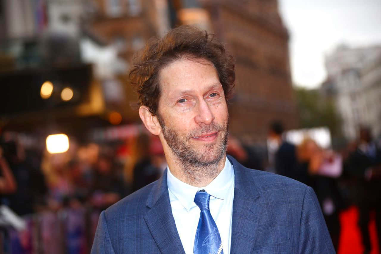 Tim Blake Nelson 桌布 壁紙 — 一個棕色捲髮、留鬍子的男人，身穿藍色格紋西裝和藍色領帶，站在紅地毯上，身後是模糊的城市建築和燈光。