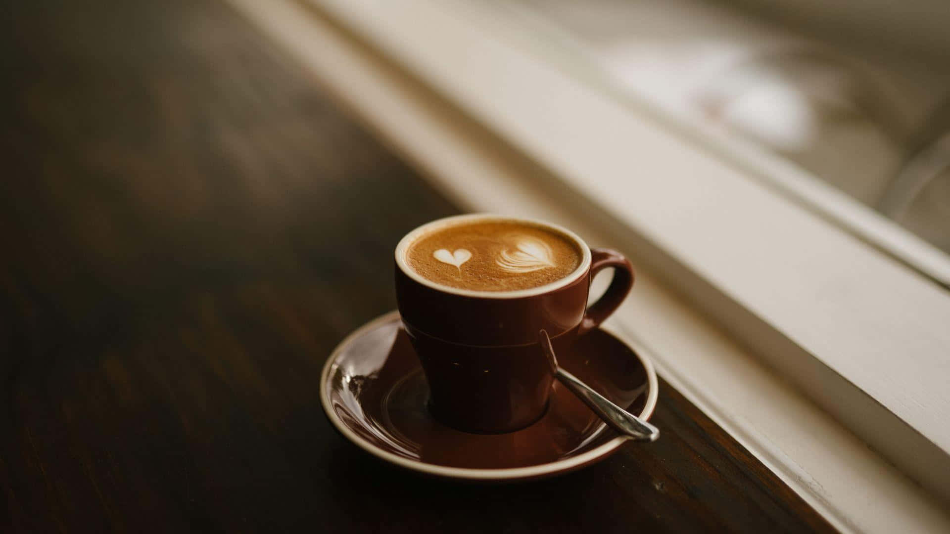 Brown Coffee 桌布 壁紙 — 一個棕色陶瓷咖啡杯，帶有心形拉花藝術，放在深色木桌上陽光照射的窗台碟子上。