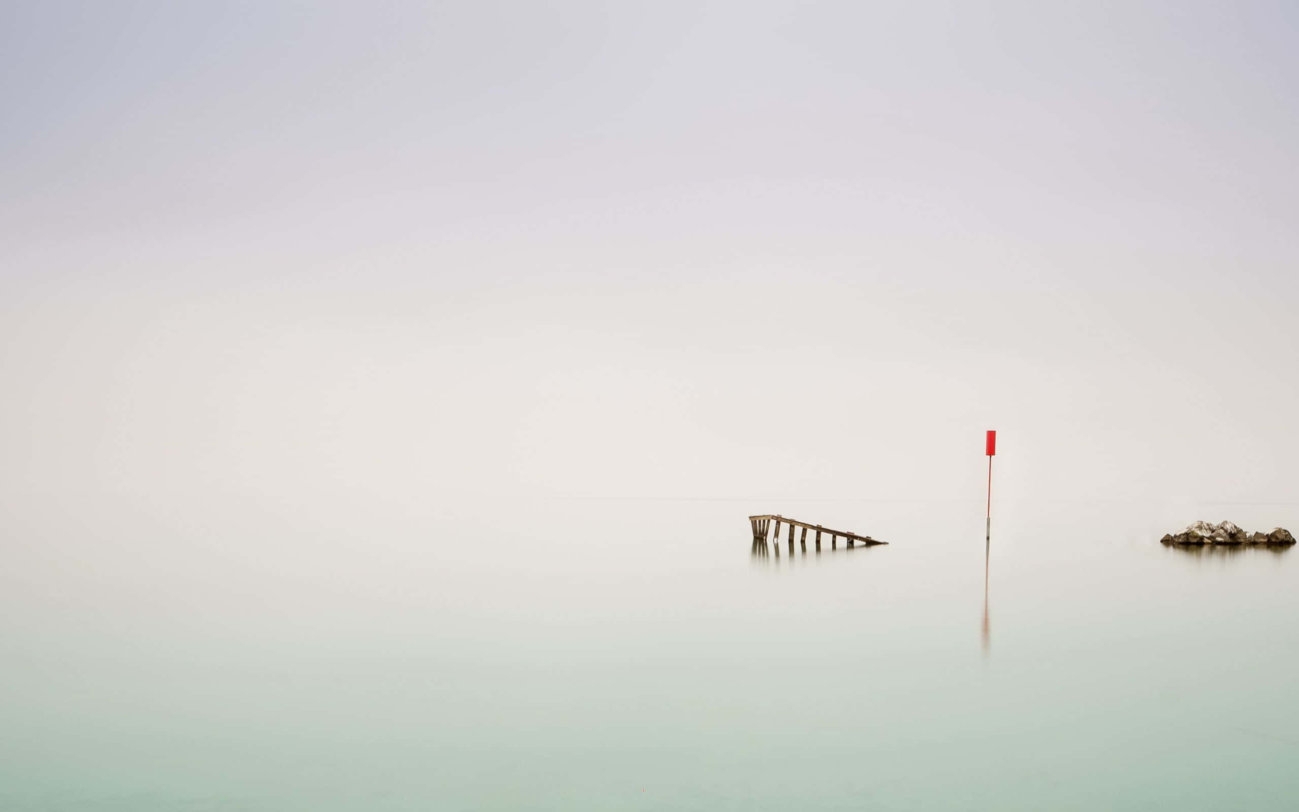 Minimalist Ocean 桌布 壁紙 — 一個極簡主義的迷霧海景，孤獨的紅色浮標和腐朽的碼頭在陰天下沉靜的灰色海水中矗立。