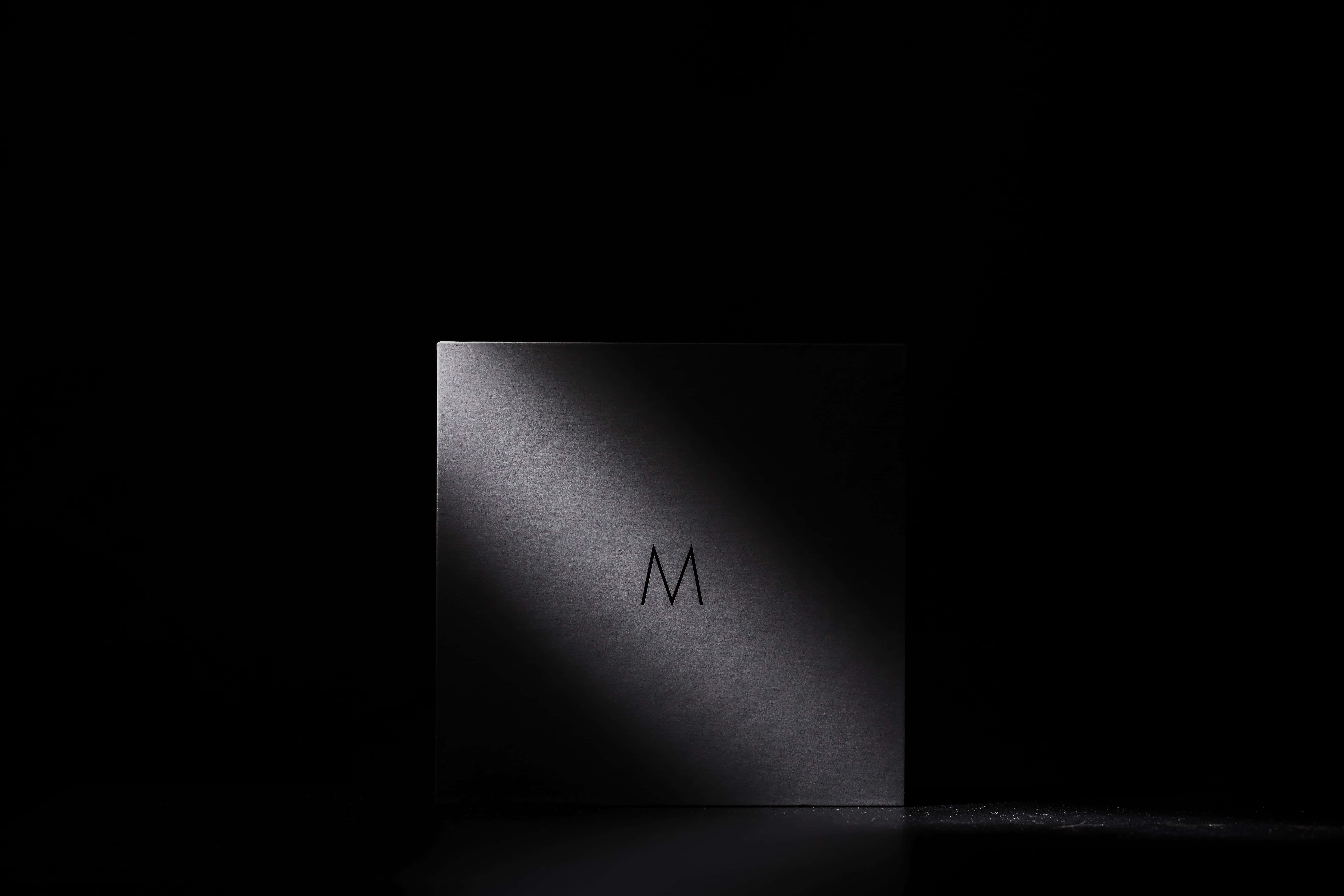 Dark Corner 桌布 壁紙 — 一個極簡的黑色正方形，帶有微妙的 'M' 標誌，由單一聚光燈照亮，背景漆黑一片。