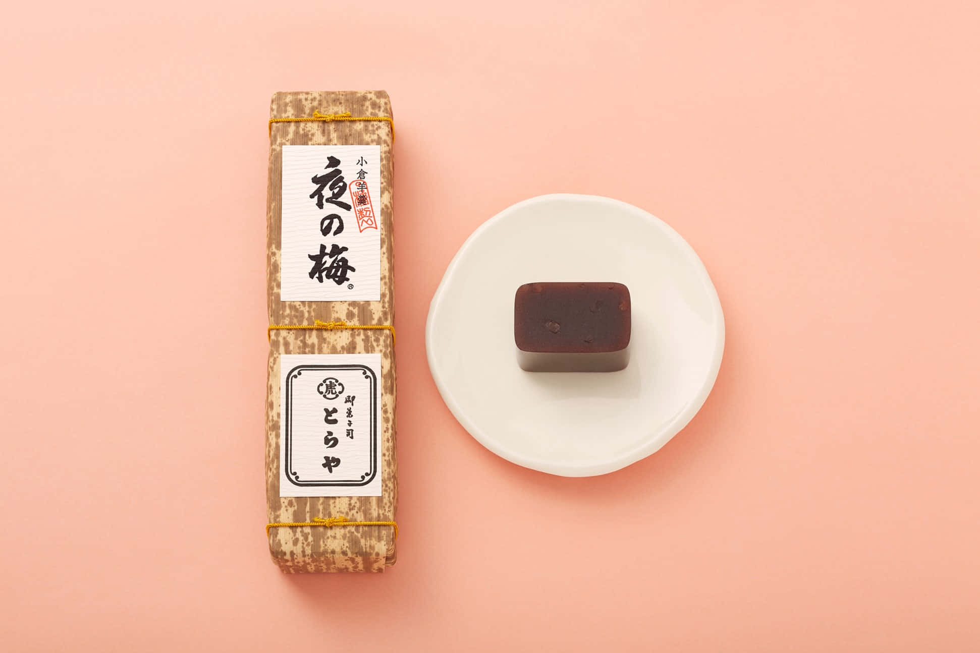 Japanese Sweets 桌布 壁紙 — 一個極簡風格的平鋪構圖，展示傳統日本梅子醬包裝和一個深棕色方形糖果放在白色碟子上，背景是柔和的粉紅色。