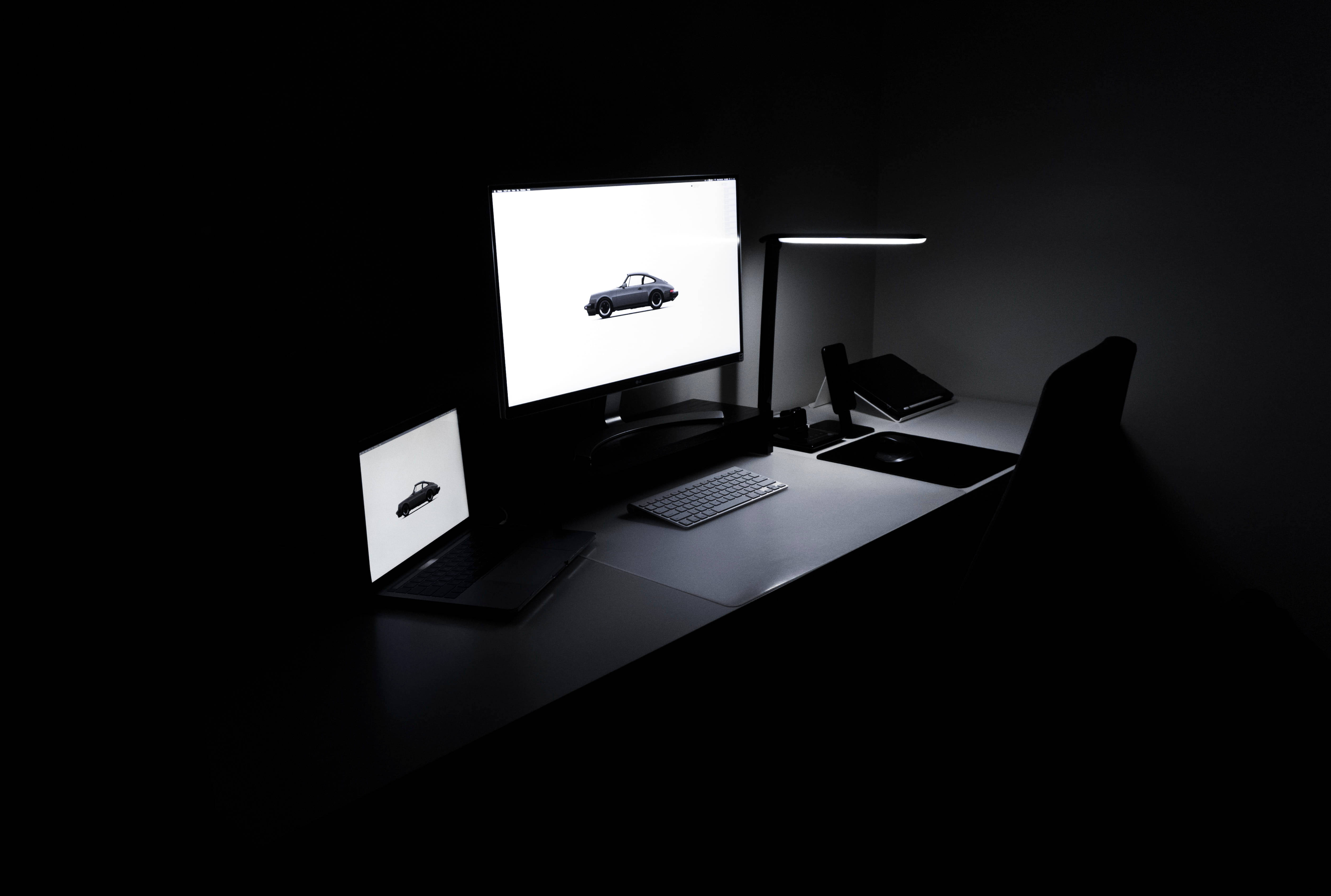 Laptop 桌布 壁紙 — 一個極簡風格的黑暗房間，配有一張時尚的書桌，由一盞燈照亮，在兩個發光的屏幕上顯示一輛經典汽車。