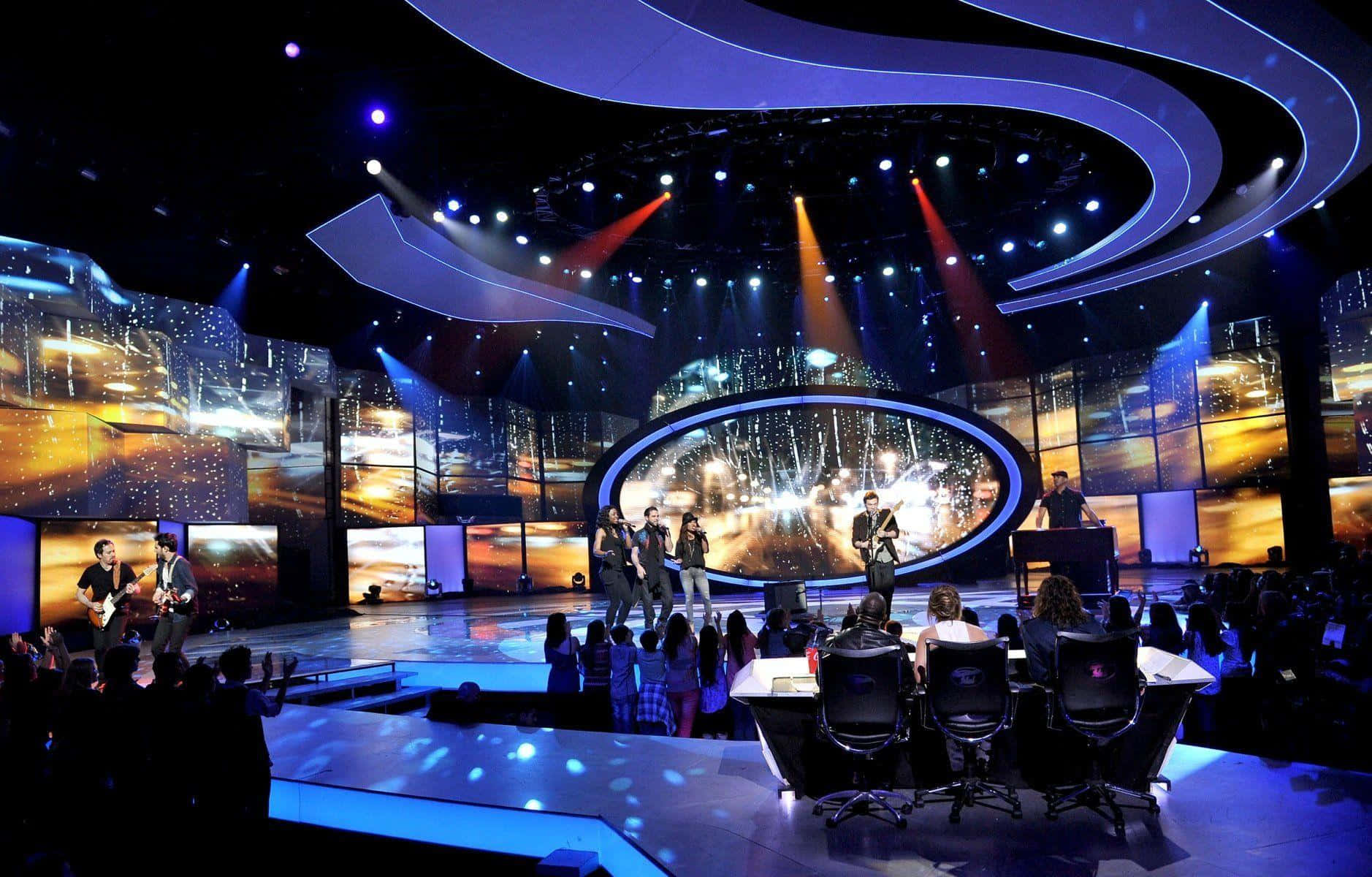 american idol 桌布 壁紙 — 一個樂隊在未來風格的舞台上表演，發光的藍色曲線和溫暖的橙色聚光燈照亮了音樂家和評判。