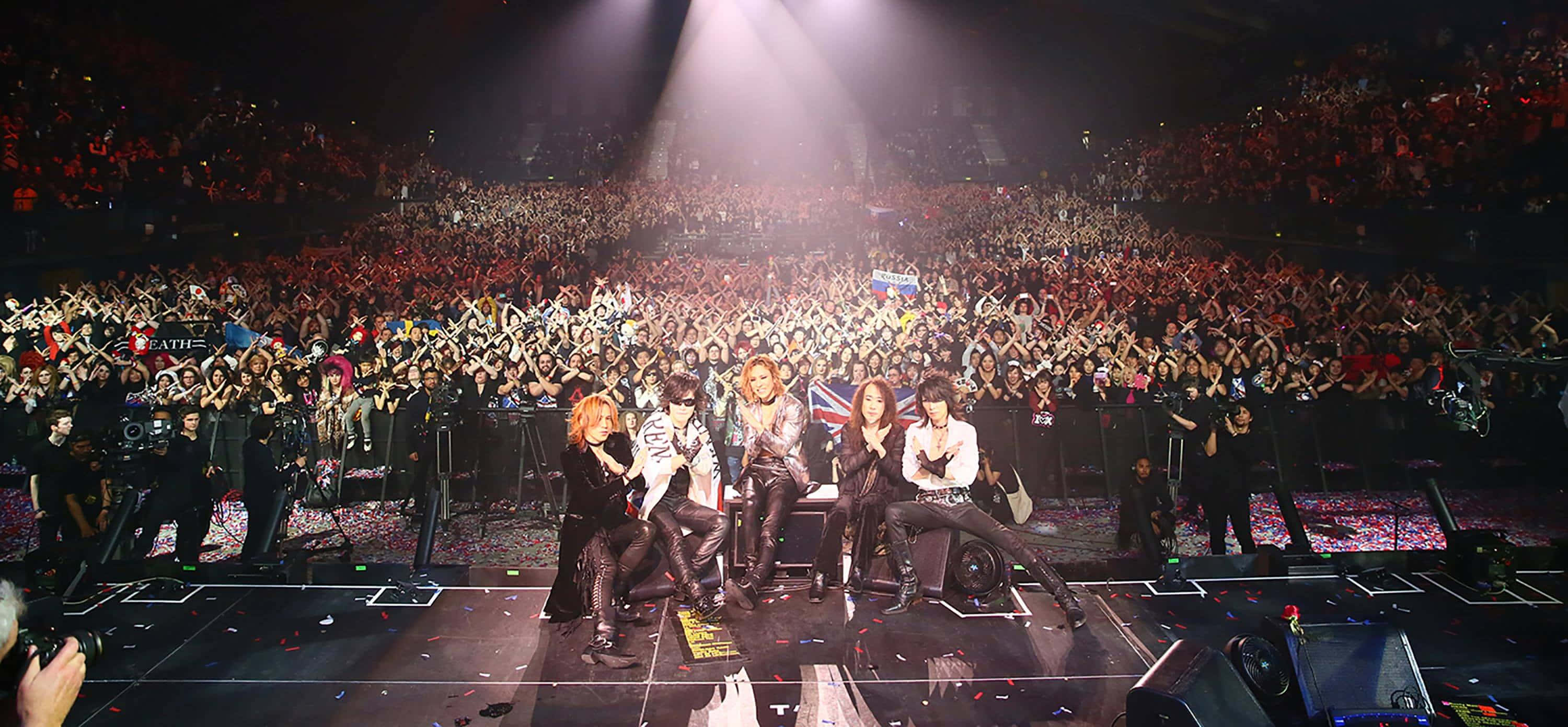 Visual Kei 桌布 壁紙 — 一個樂隊在燈光璀璨的舞台上表演，面對著擠滿場館、揮舞著螢光棒的龐大群眾。