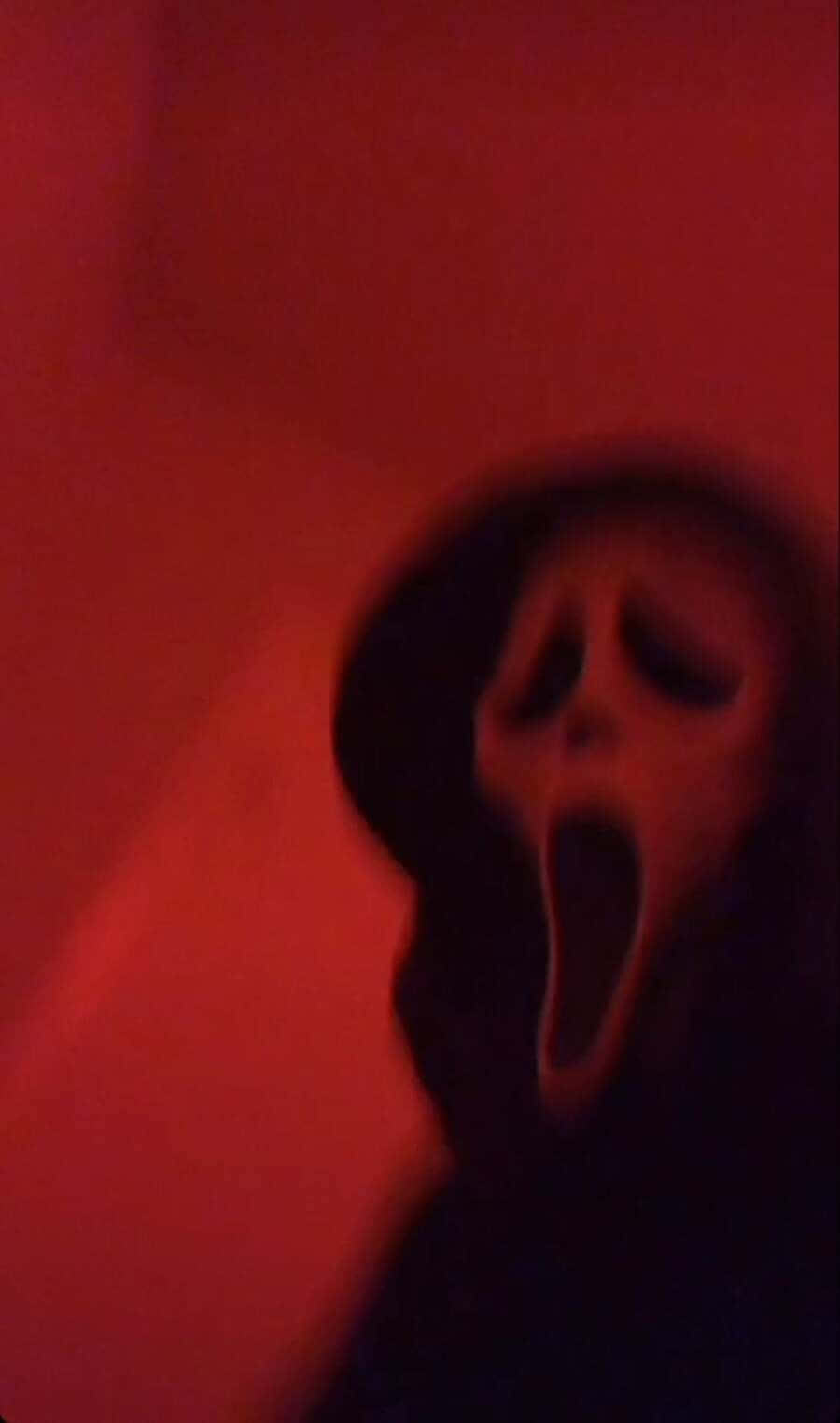 Ghostface Aesthetic 桌布 壁紙 — 一個模糊的鬼影戴着白色尖叫面具，在陰暗的紅光中站在黑暗的牆壁前。