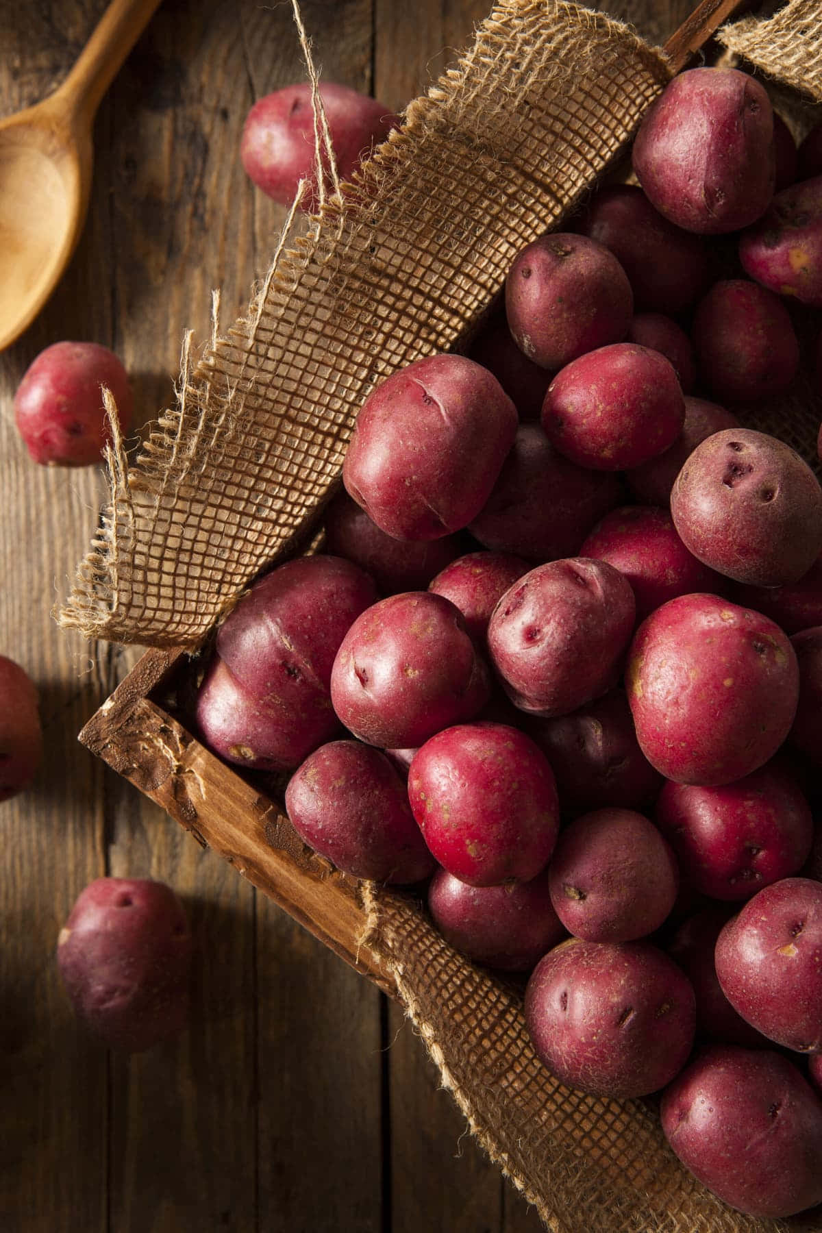 Red Potatoes 桌布 壁紙 — 一個樸素的木籃子裝滿了鮮紅色的馬鈴薯，放在一張風化的木桌上，旁邊有一把木勺。