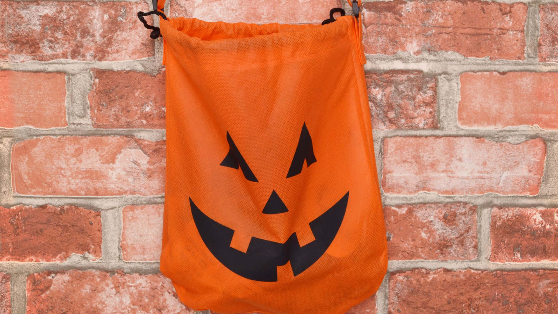 Trick Or Treat Bags 桌布 壁紙 — 一個橙色嘅萬聖節trick-or-treat袋，上面有黑色嘅jack-o'-lantern面，掛喺紅磚牆上。